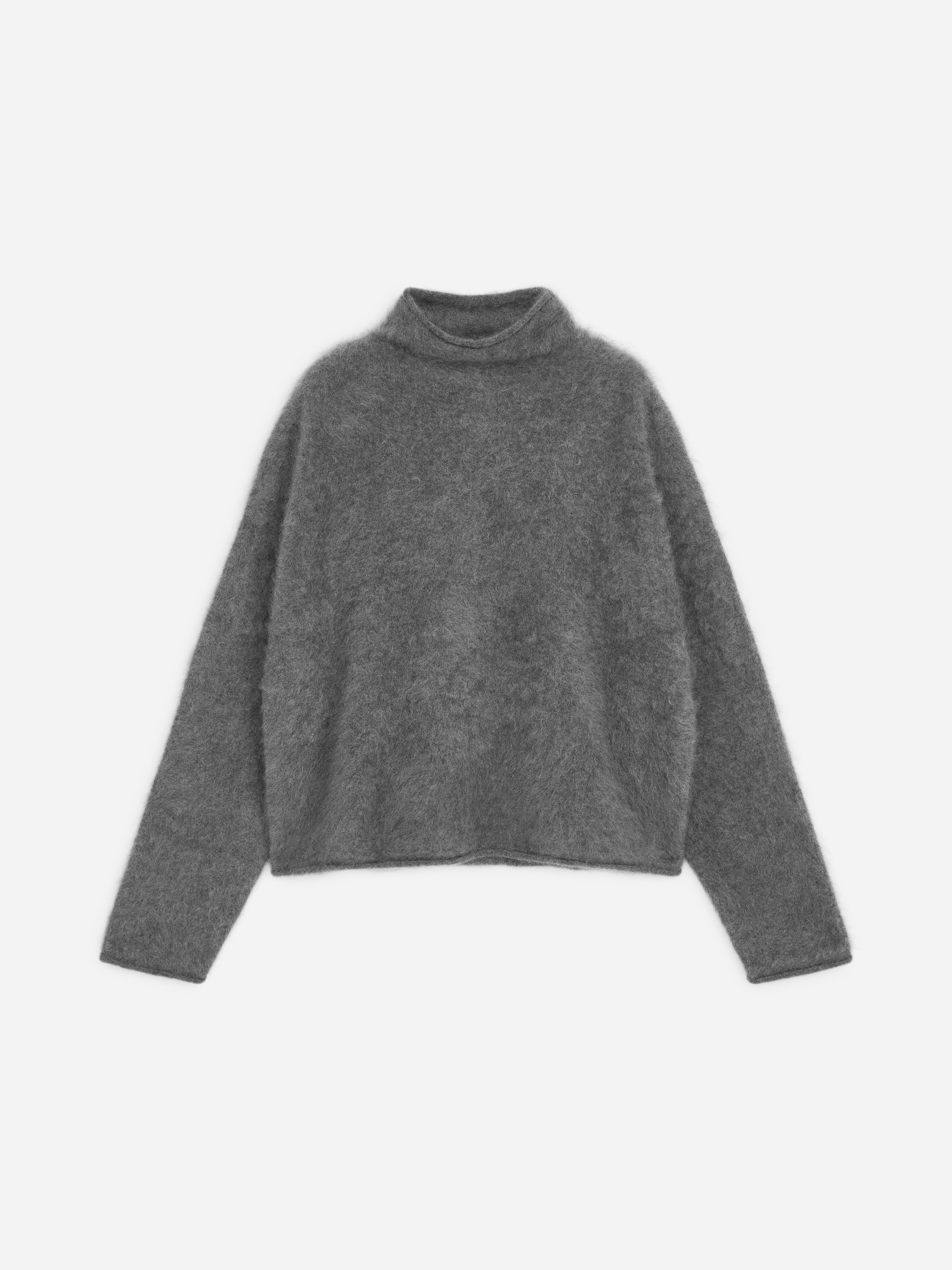En grÃ¥, strukturert strikkegenser med rullekrage bÃ¦res av modellen, med en avslappet silhuett, lange ermer og hÃ¸y hals med rullede kanter. Genseren er lÃ¸st stukket ned i linningen pÃ¥ svarte denimplagg, festet med et tynt svart belte.