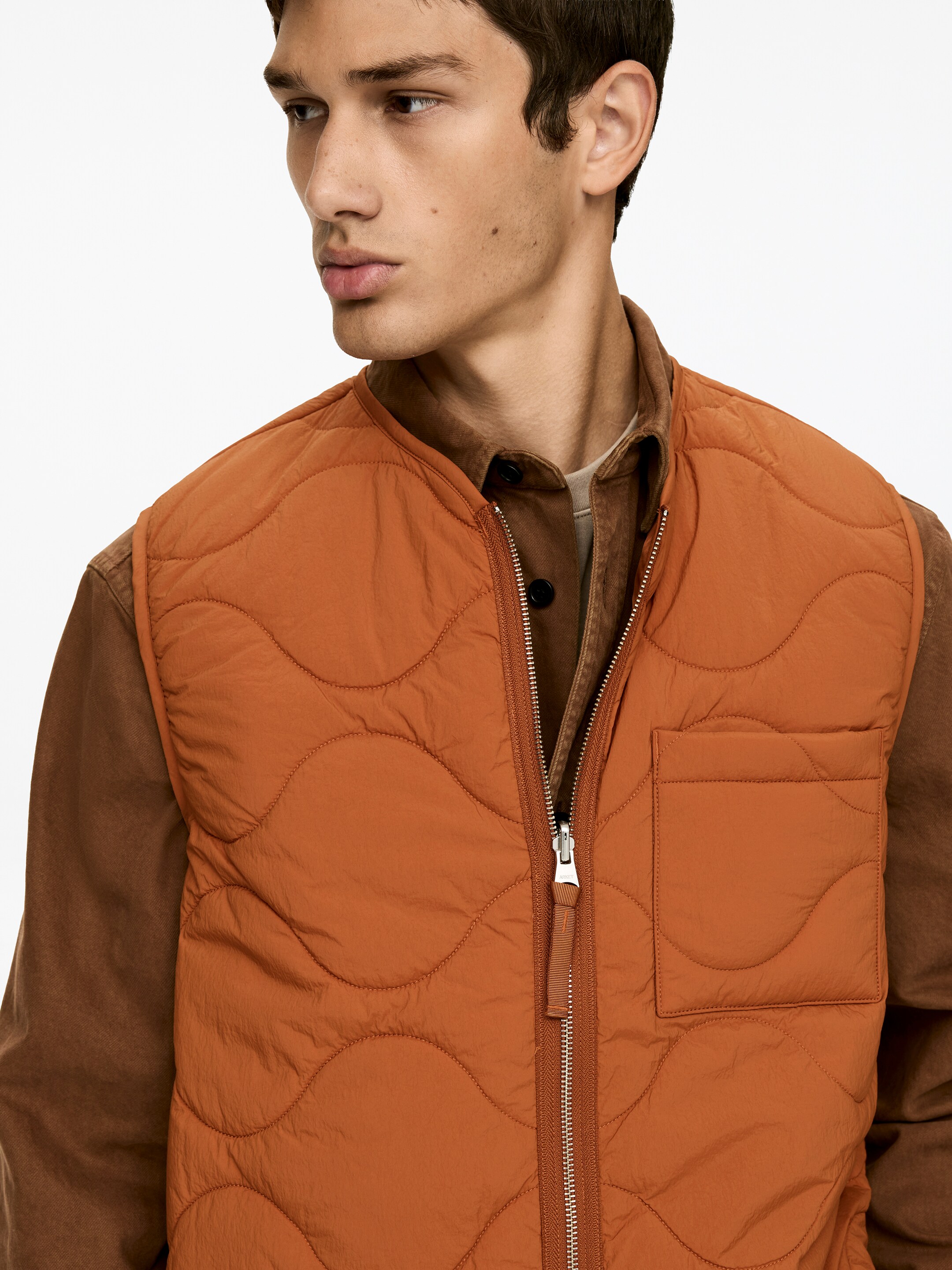 Agrandir l'image: Un gilet matelassé orange est présenté de face, porté par-dessus une chemise marron à col. Il présente un motif matelassé ondulé, une fermeture éclair en métal de longueur totale avec une tirette en tissu, ainsi qu'une poche plaquée sur la poitrine gauche du porteur.