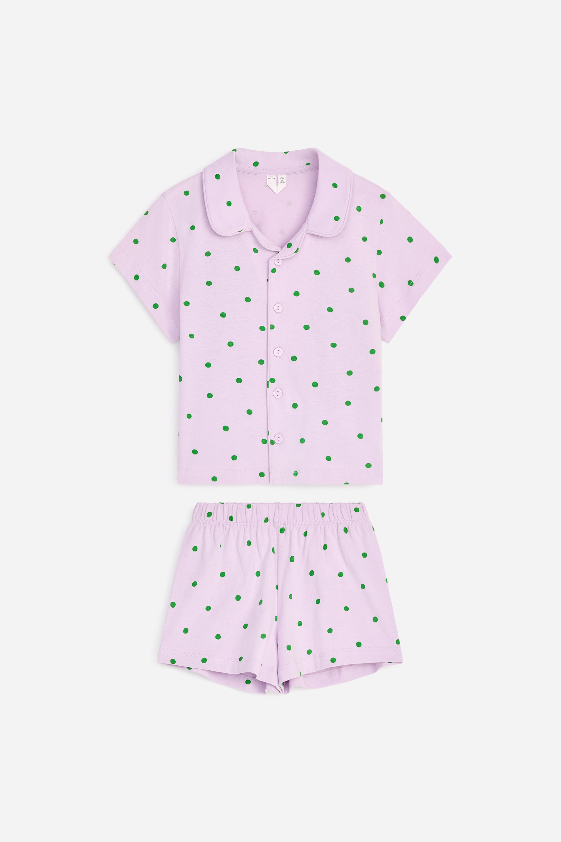 Se større billede: Et lyse lilla pyjamassæt vises forfra og har grønne polkaprikker. Den kortærmede top har en rund krave og en knaplukning foran, og de matchende shorts har en elastisk linning.