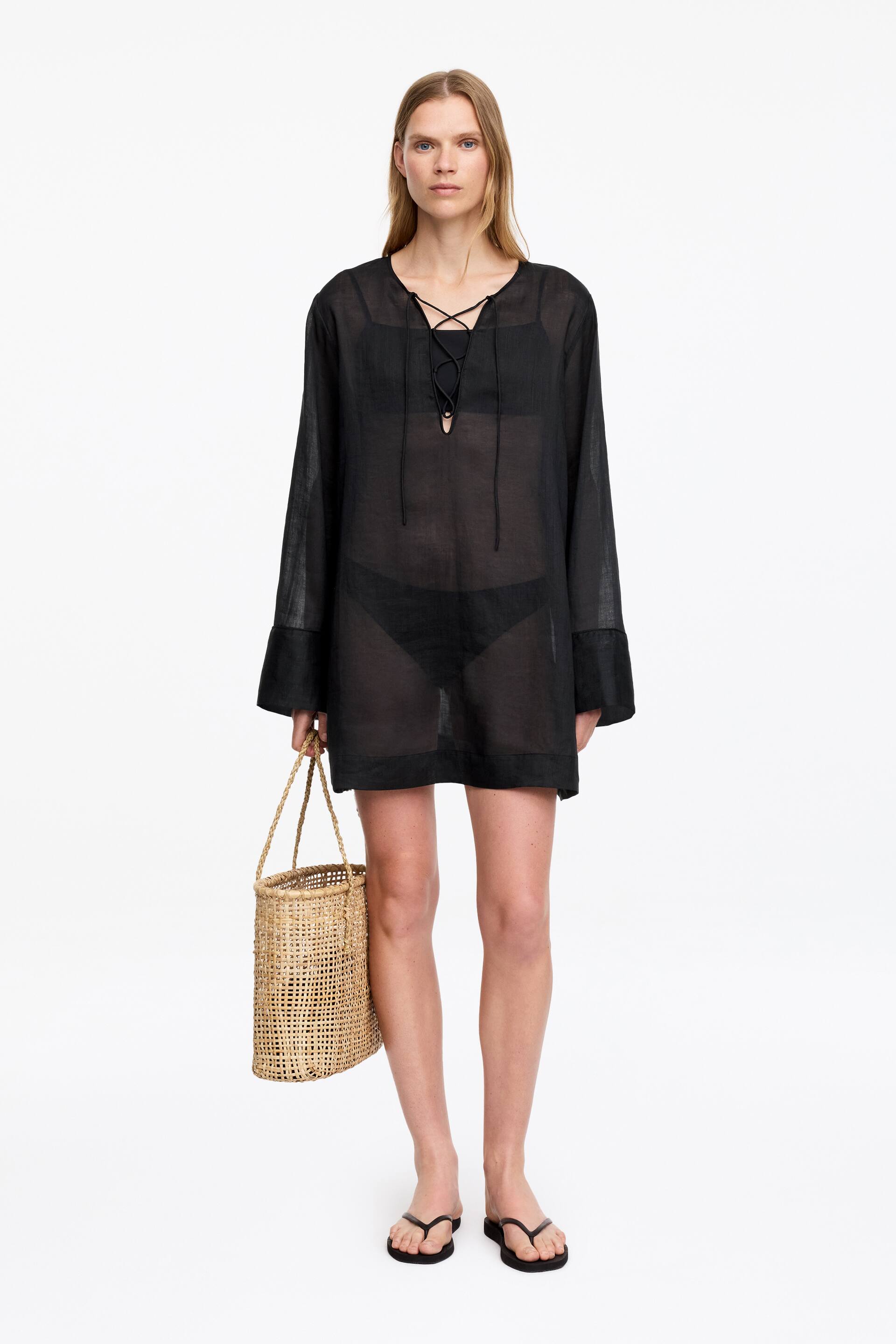 Nagyobb kép megtekintése: A fekete áttetsző strandruha V-nyakkivágással rendelkezik, fűzős részlettel és hosszú, bő ujjakkal, széles mandzsettával. A térd fölé esik, és alatta látható a fekete fürdőruha.