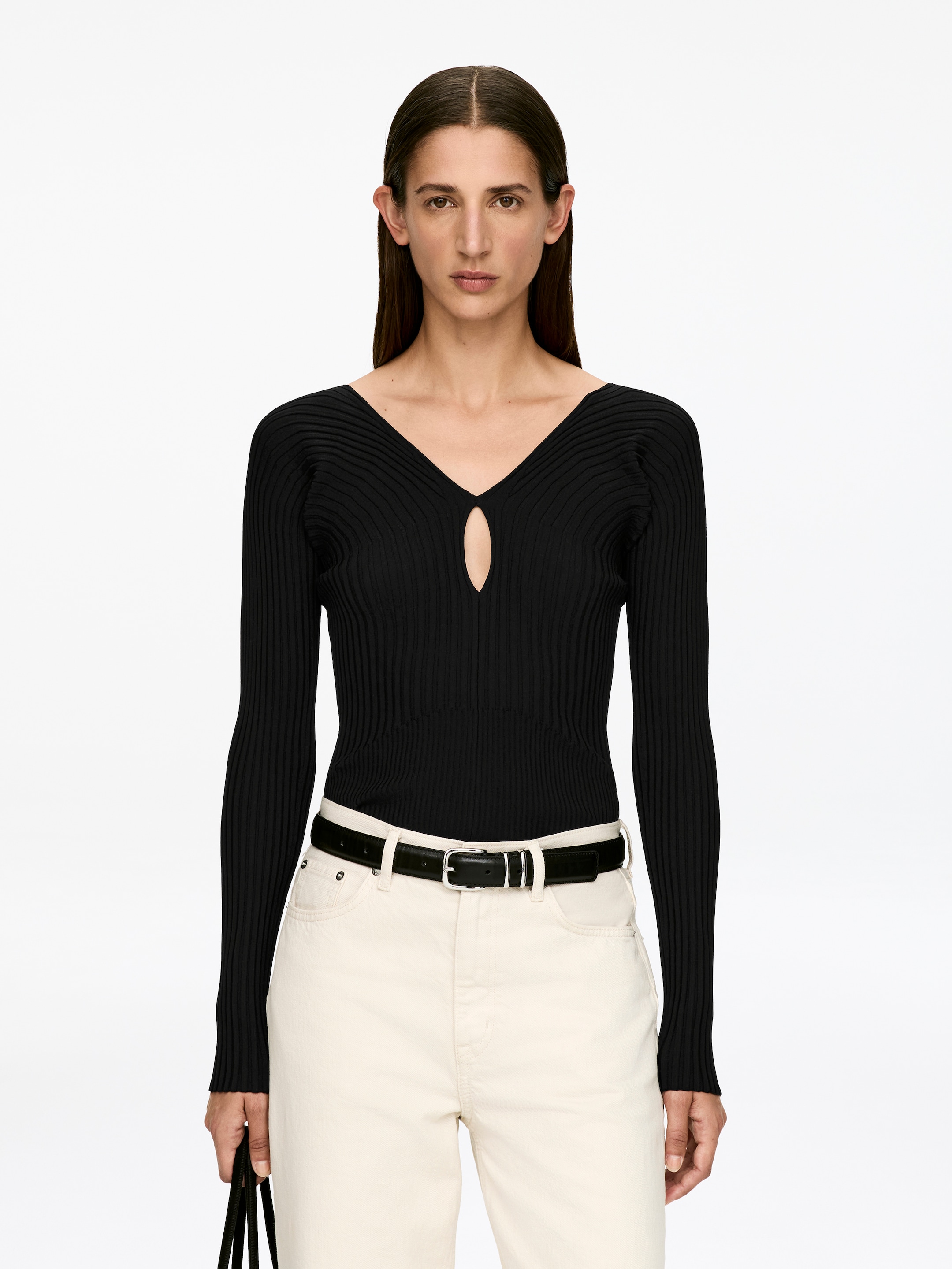 Grotere afbeelding bekijken: Een zwart ribgebreid topje met een sleutelgatopening aan de voorkant en een diepe V-hals wordt gedragen door het model. Het slim-fit topje, voorzien van lange mouwen, is ingestopt in lichtgekleurde broeken met een zwarte riem.