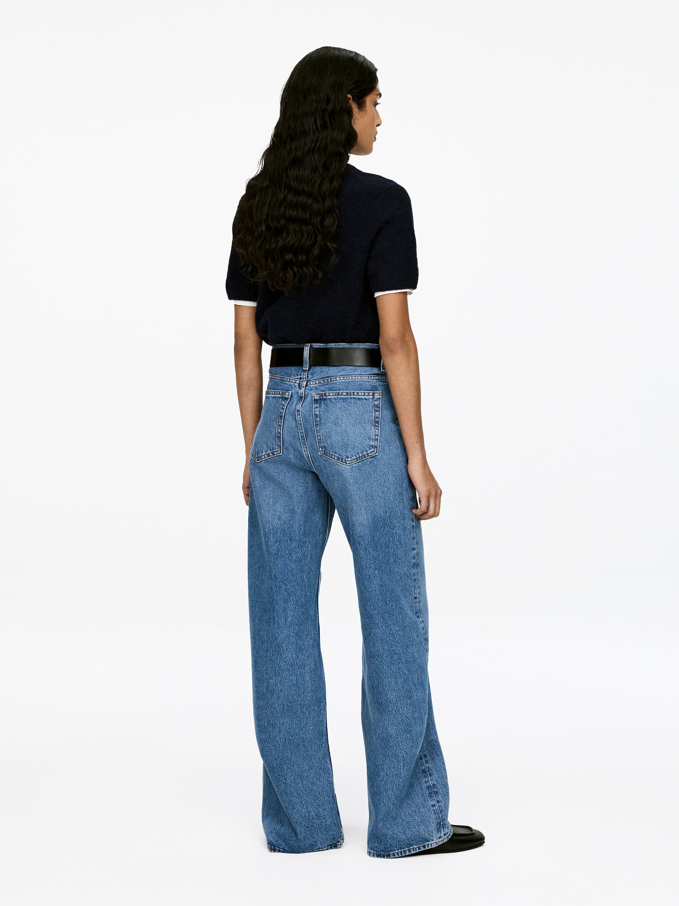 Visa större bild: LUPINE-jeans bärs av modellen, och de har en hög midja, smal passform och utsvängda ben i ljus tvätt denim, med synliga fickor bak. Ett svart bälte drar åt midjan.