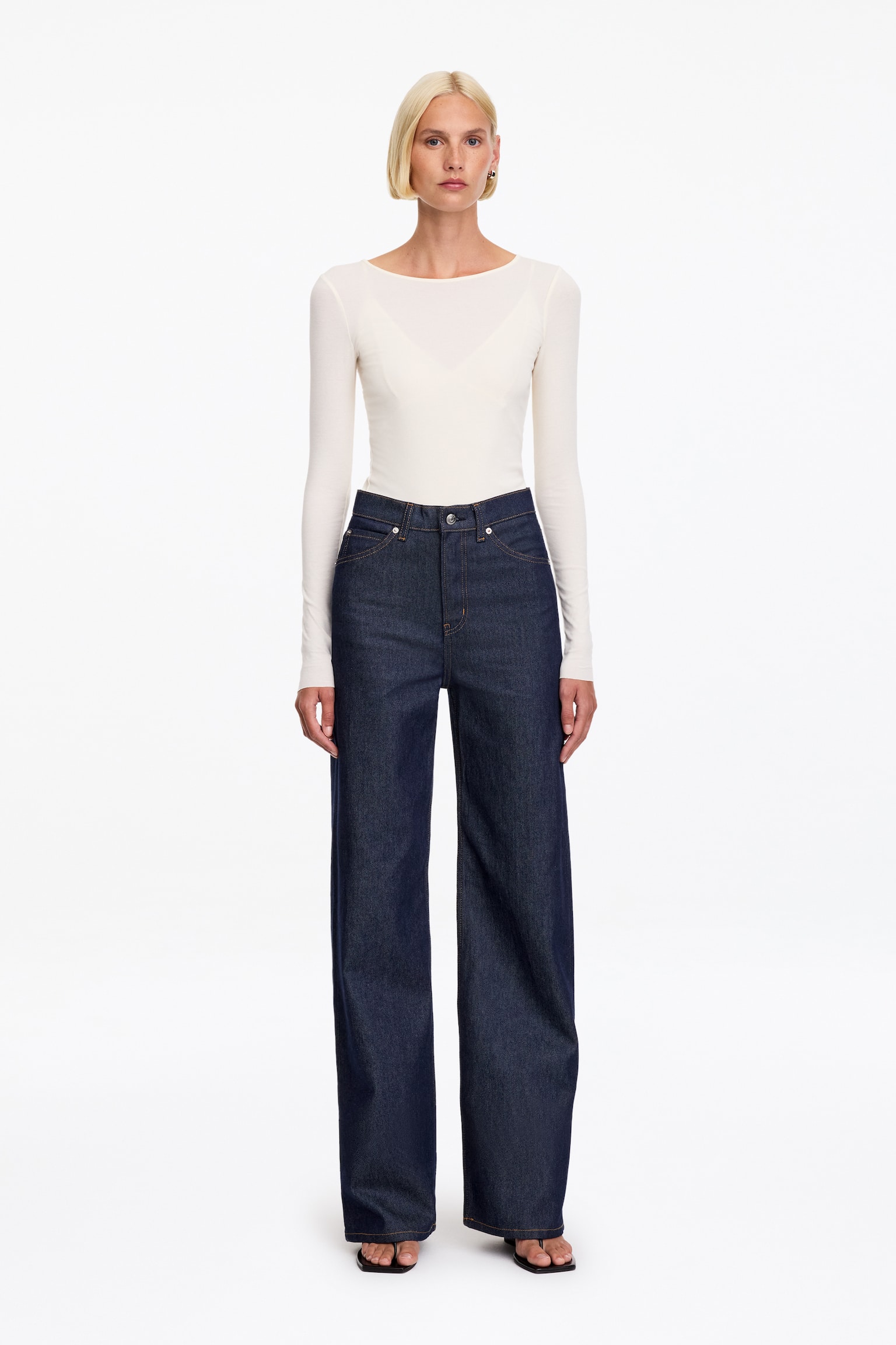 Een paar donkerblauwe denim jeans wordt gepresenteerd vanaf de VOORKANT, met een hoge taille, een ritssluiting en voorzakken met klinknagels. De jeans is ontworpen met een slanke pasvorm die uitloopt in wijde pijpen, en ze zijn gedetailleerd met zichtbare contrasterende stiksels. - 1