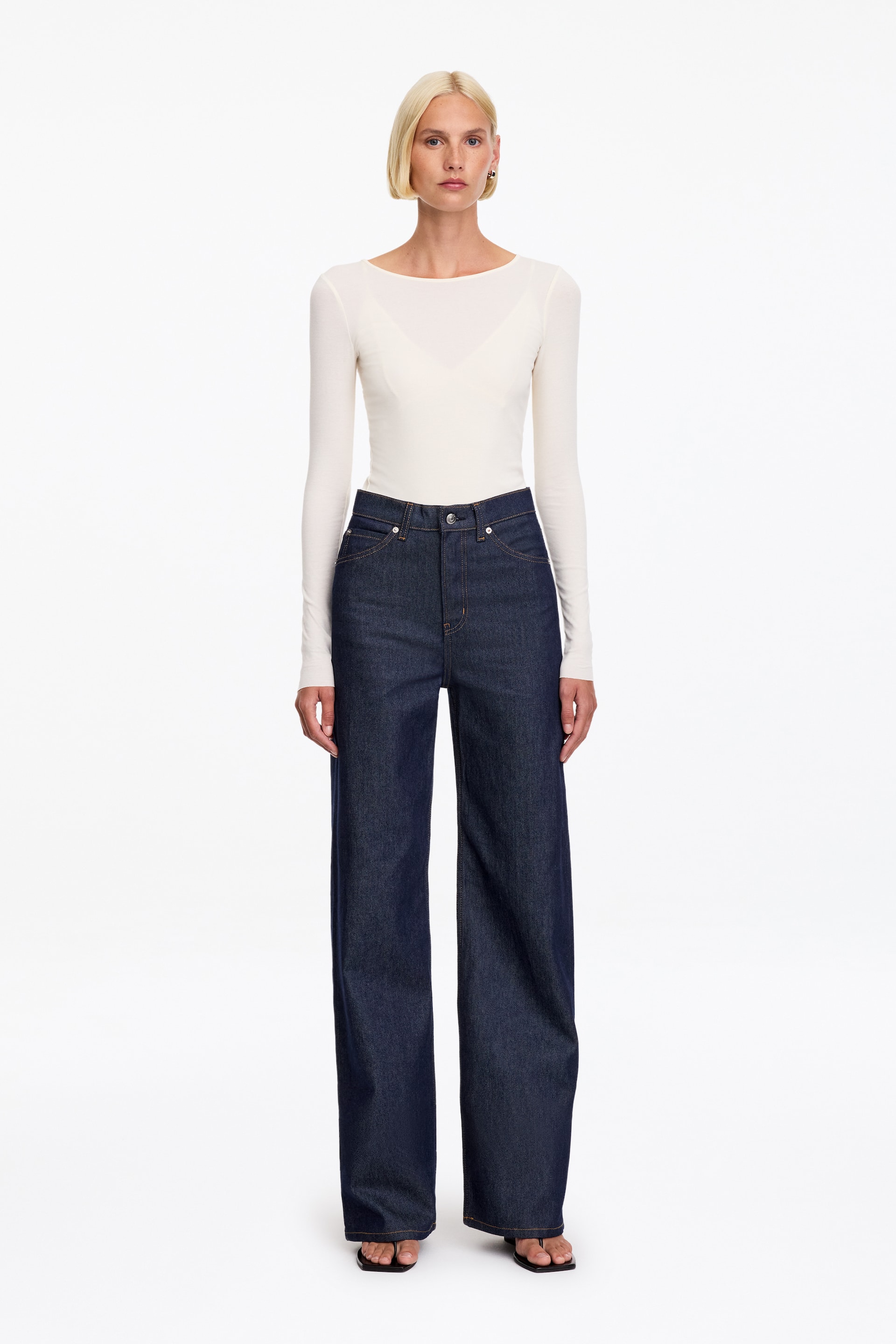 Ver imagem maior: Jeans de ganga azul-escura, com cintura subida e pernas à boca de sino, usados pelo modelo. As calças de ganga são combinadas com um top creme justo de manga comprida, metido na cintura.