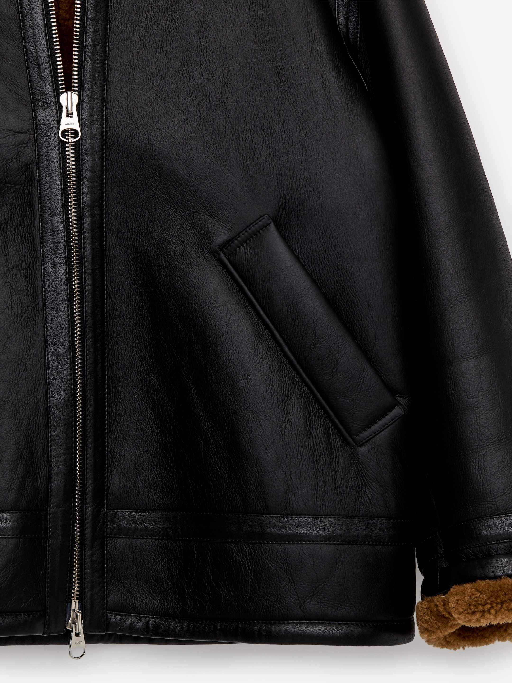 Agrandir l'image: Une veste en shearling surdimensionnée présente un extérieur en cuir noir avec une doublure en fourrure marron, visible le long de la fermeture éclair argentée à double sens partiellement ouverte qui descend le devant et au poignet de la manche. Une poche passepoilée en biais est positionnée sur le devant de la veste.