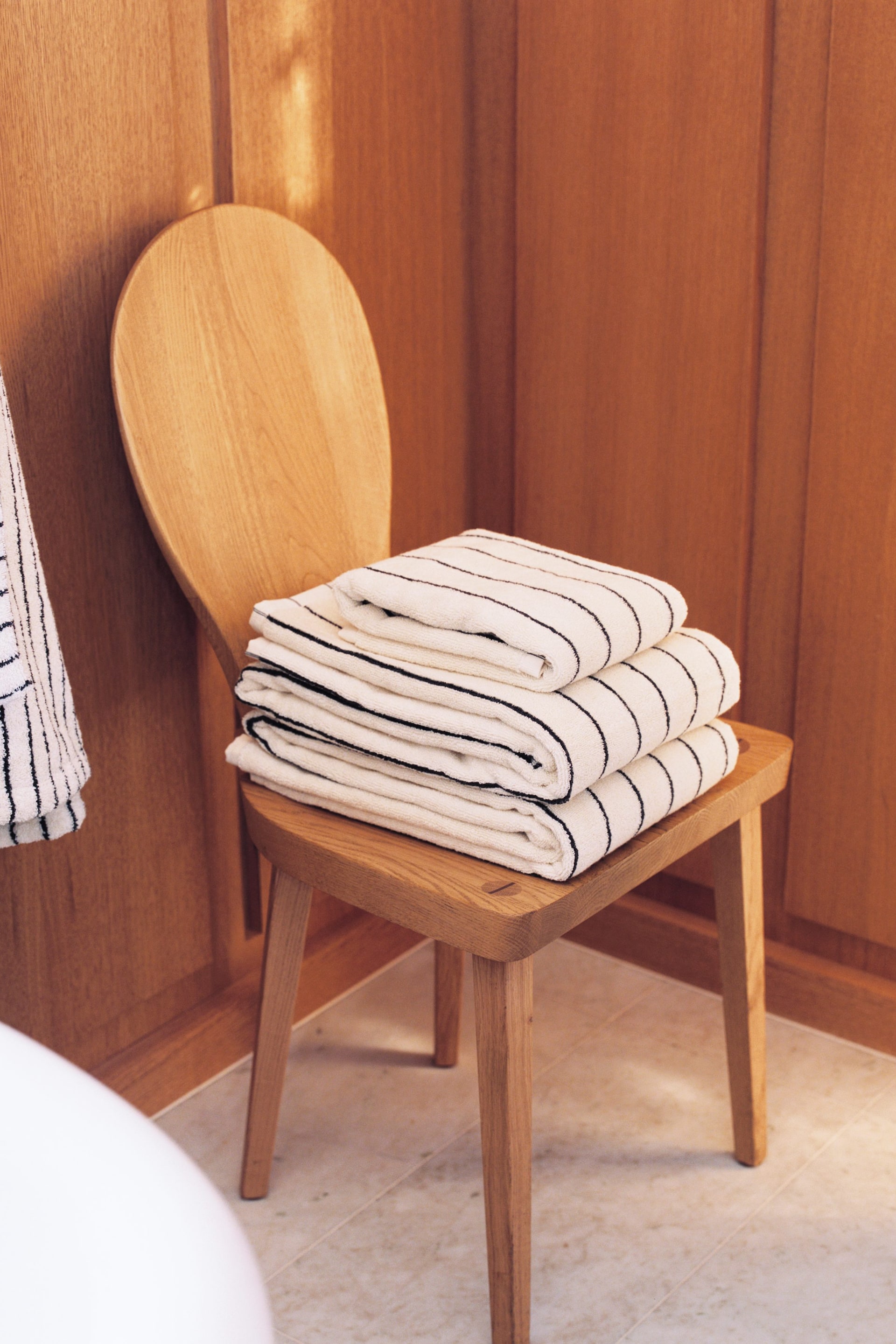 Agrandir l'image: Trois serviettes de couleur crème, ornées de fines rayures horizontales noires, sont soigneusement pliées et empilées sur une chaise en bois. Une serviette similaire à rayures est partiellement visible, suspendue à gauche.