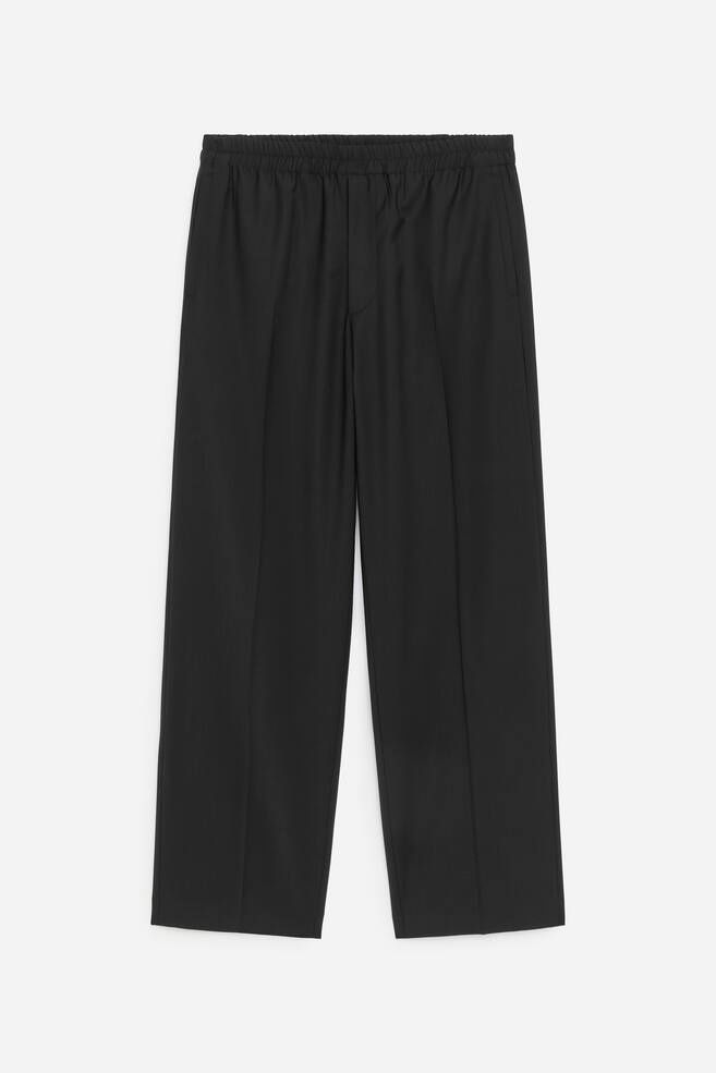 Zwarte broek wordt van voren weergegeven, met een elastische tailleband en subtiele zijzakken. De broek heeft een rechte, wijde pijp met opvallende verticale vouwen die over de voorkant van elke pijp lopen. - 1