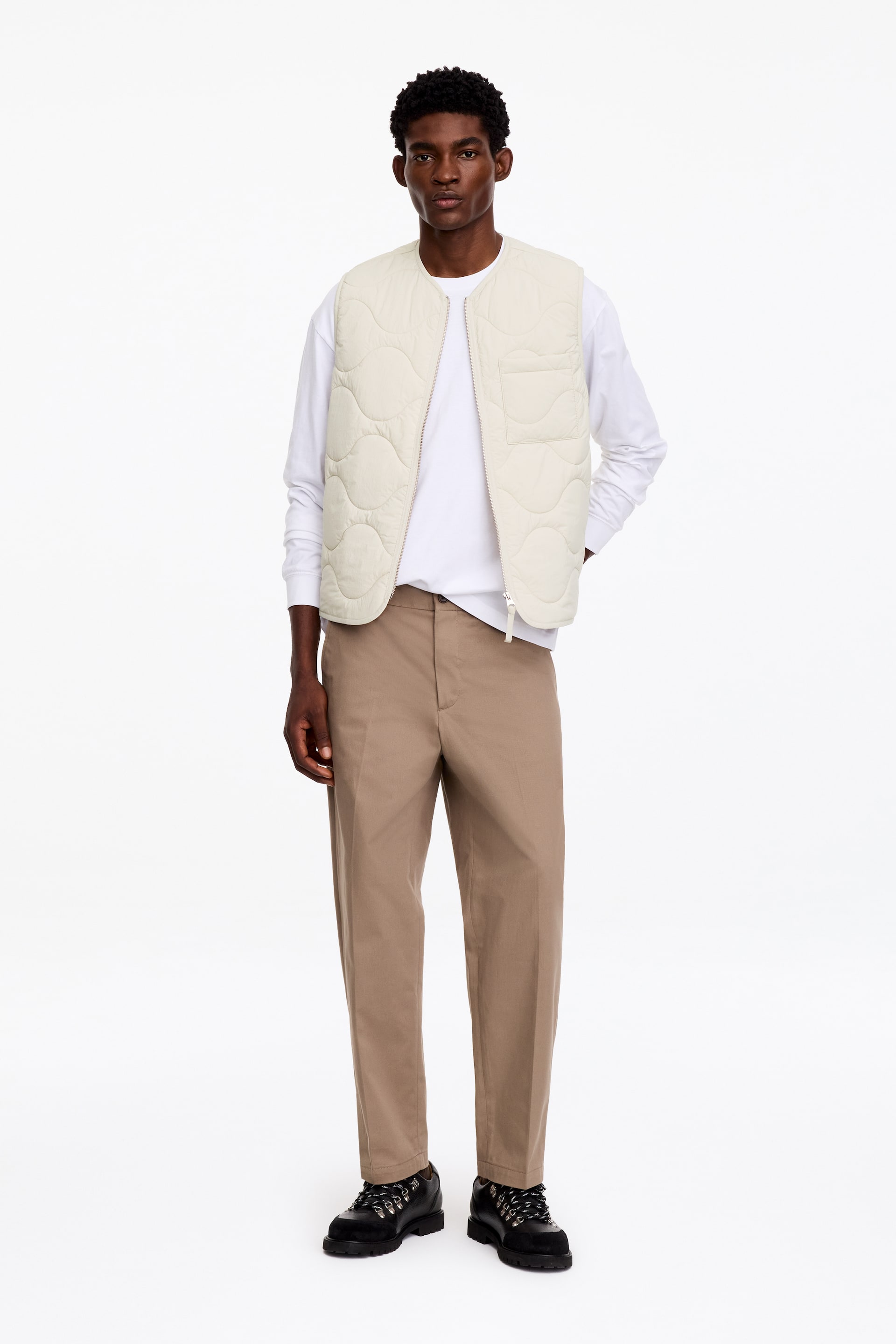 View larger image: The model is wearing an off-white quilted gilet with an onion-pattern design and a single chest pocket, worn open over a white long-sleeved T-shirt. The gilet is styled with beige straight-leg trousers and black lace-up boots.