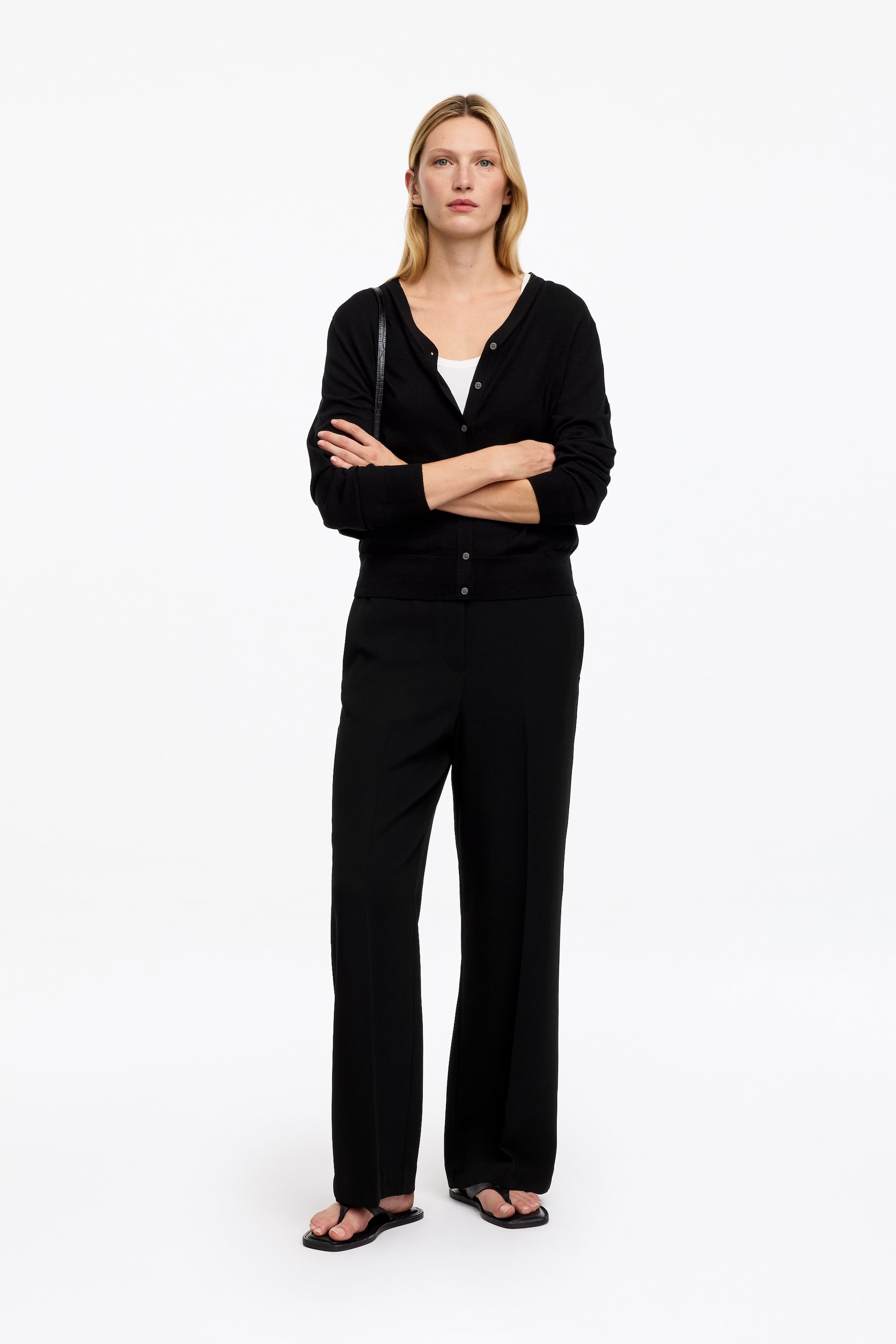 Visualizza immagine più grande: La modella indossa pantaloni neri con silhouette a gamba dritta e piega stirata sul davanti. I pantaloni sono abbinati a un cardigan nero con bottoni, un top bianco e sandali neri bassi.
