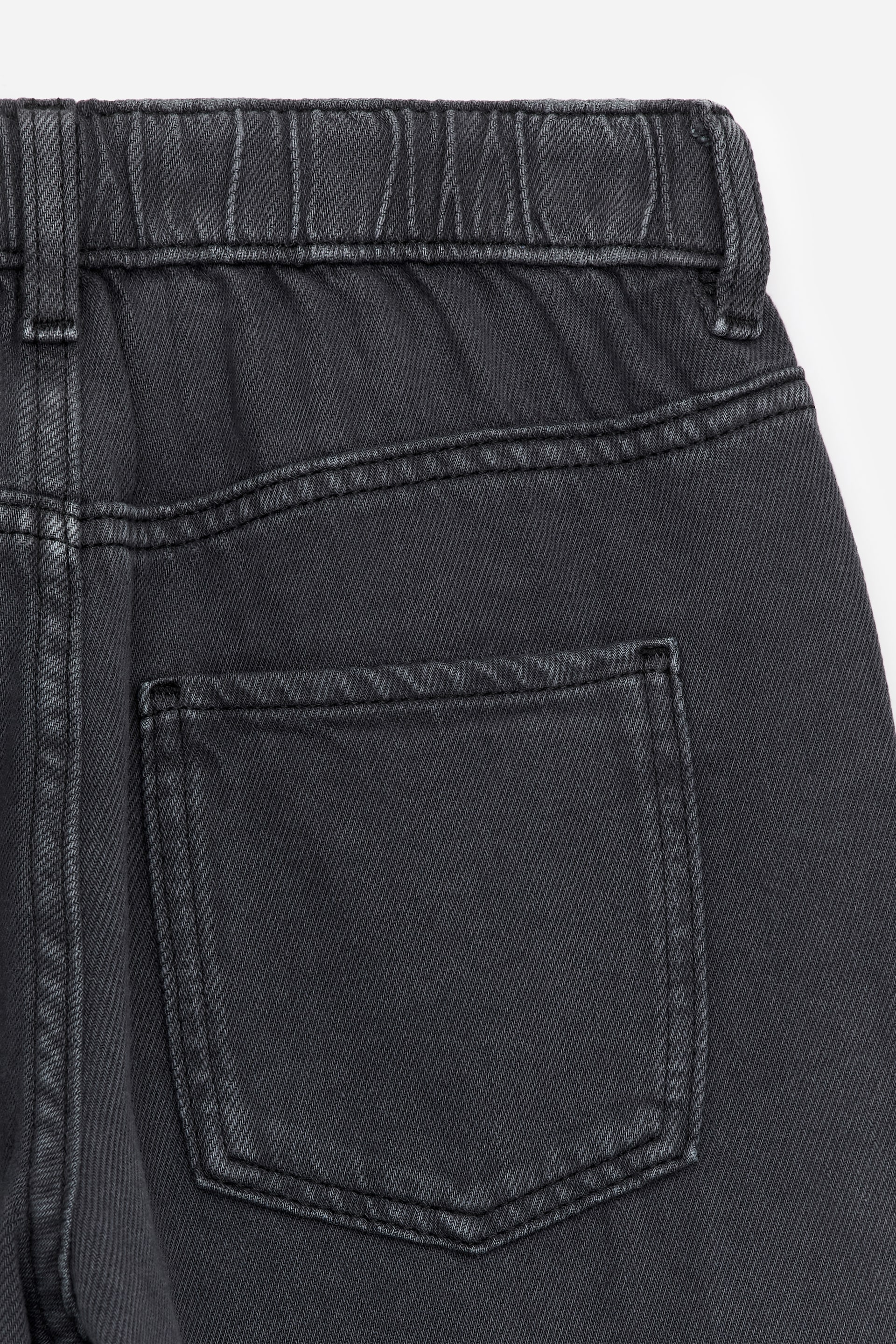 Ver imagem maior: A parte de trás das calças denim cinzento-escuro apresenta um cós elástico franzido. Um único bolso aplicado é visível na parte de trás, e o tecido denim exibe costuras proeminentes.