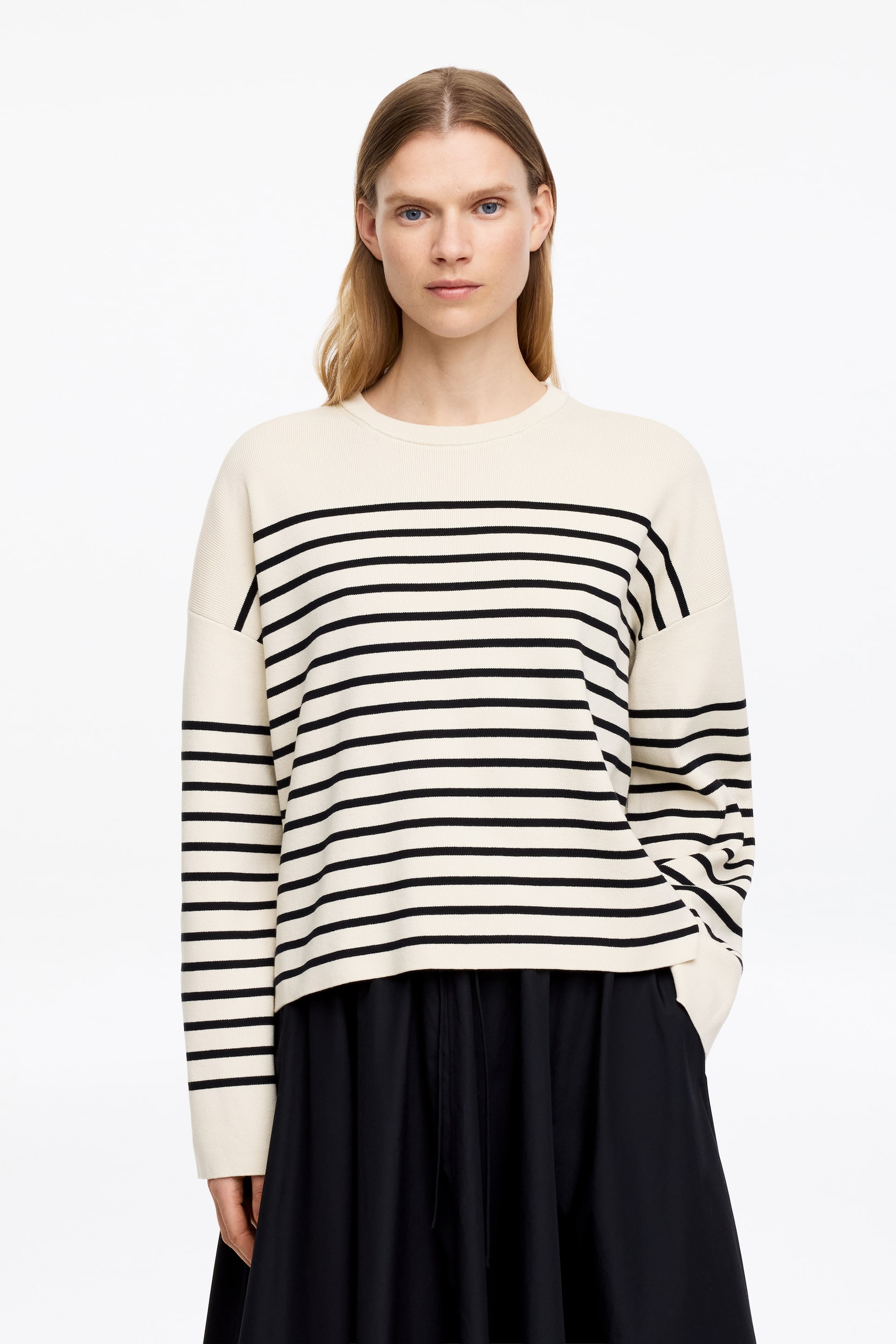 Zobacz większy obraz: Modelka ma na sobie kremowy sweter w czarne poziome paski, z okrągłym dekoltem, opadającymi ramionami i francuskim krojem ramion. Ten sweter o regularnym kroju posiada boczne rozcięcia, a modelka wkłada jedną rękę do kieszeni czarnych spodni lub spódnicy.