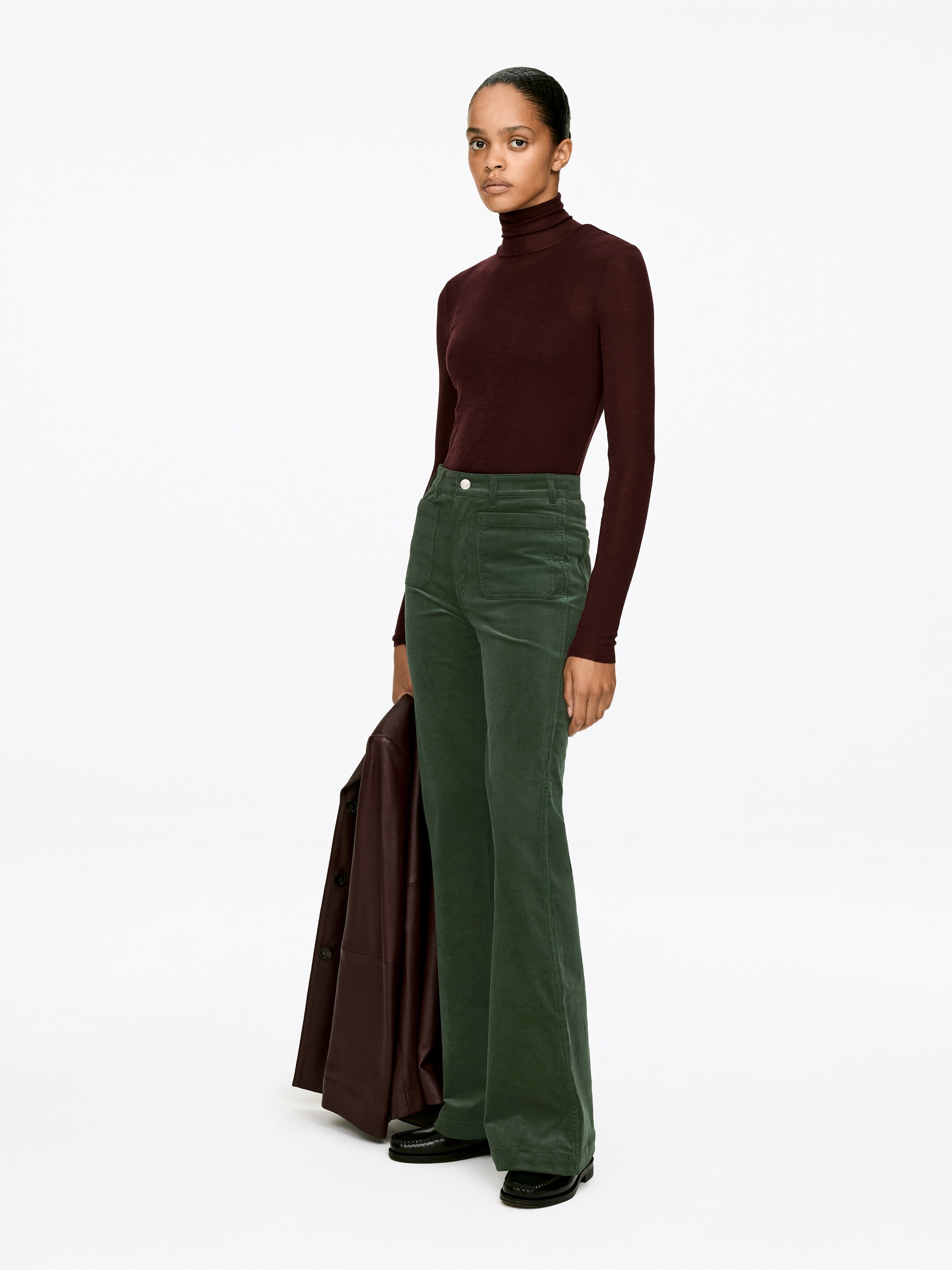 Ver imagen más grande: Una persona está presentando una vista de tres cuartos, modelando un top de cuello alto ajustado de color burdeos oscuro combinado con unos pantalones de pana verde de talle alto y acampanados. Los pantalones presentan dos bolsillos delanteros con botones de color claro. Una chaqueta marrón oscuro se sostiene sobre un brazo, y unos mocasines negros son visibles en los pies.