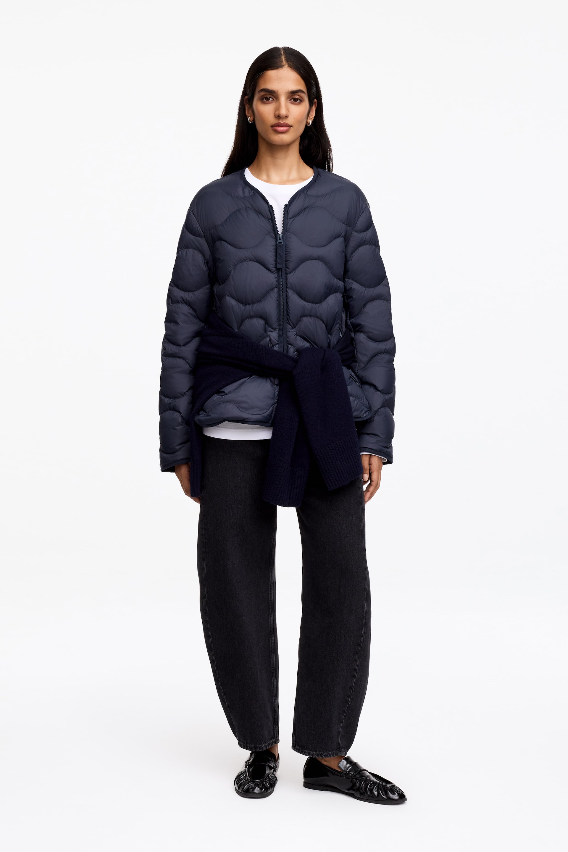 Agrandir l'image: Une veste matelassée bleu marine est présentée de face, avec un col rond sans col et une fermeture éclair frontale de couleur argentée. Elle met en valeur un motif de matelassage en forme de coquille et est portée par-dessus un t-shirt blanc, avec un pull bleu marine noué autour de la taille.