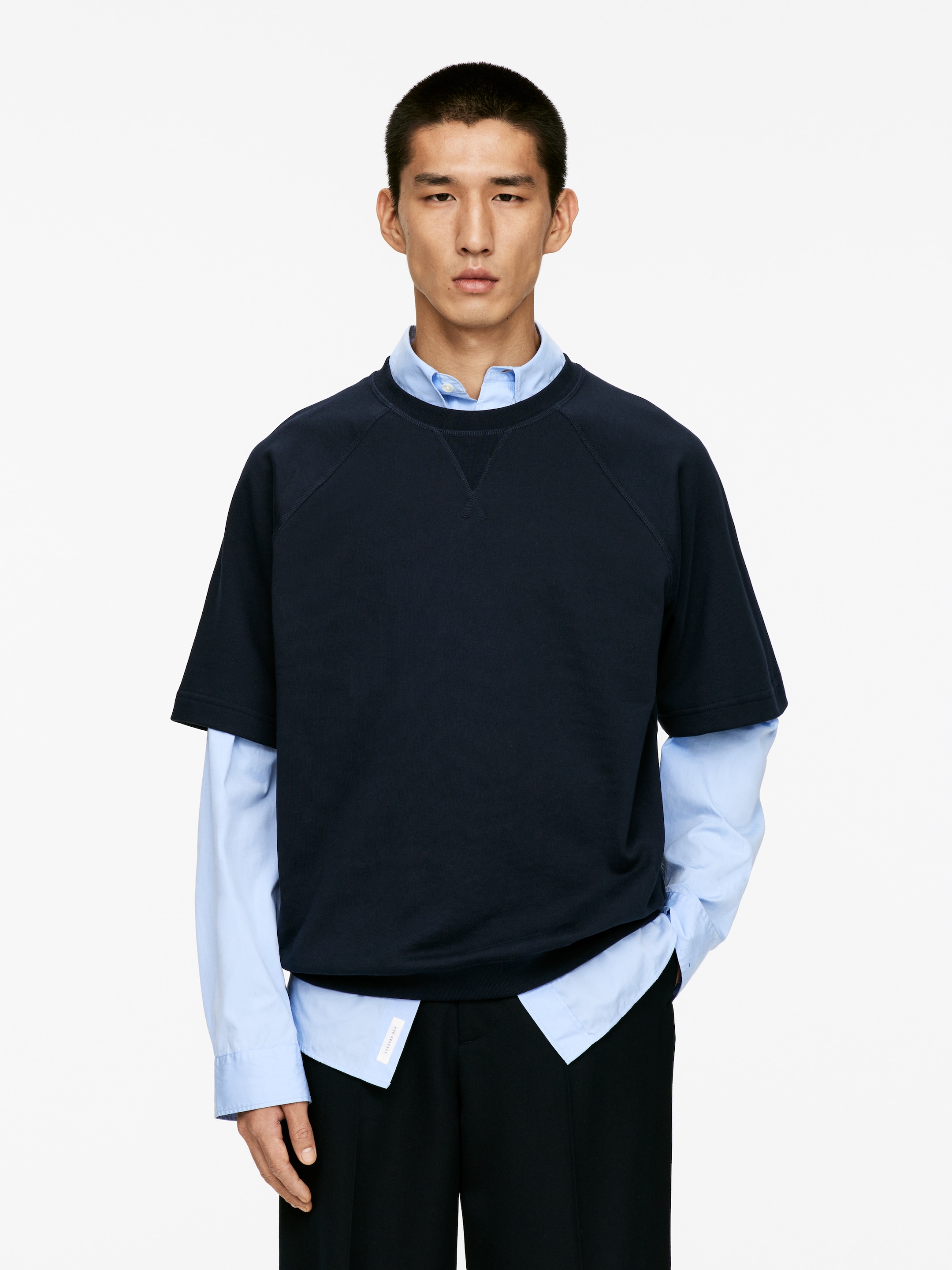 Größeres Bild anzeigen: Ein marineblaues Sweatshirt mit kurzen Ärmeln wird aus einer Frontalansicht präsentiert. Es ist mit einem Rundhalsausschnitt mit V-Stich-Detail und Raglanärmeln gestaltet und wird über einem hellblauen langärmeligen Hemd mit Kragen getragen, was eine entspannte Silhouette zeigt.