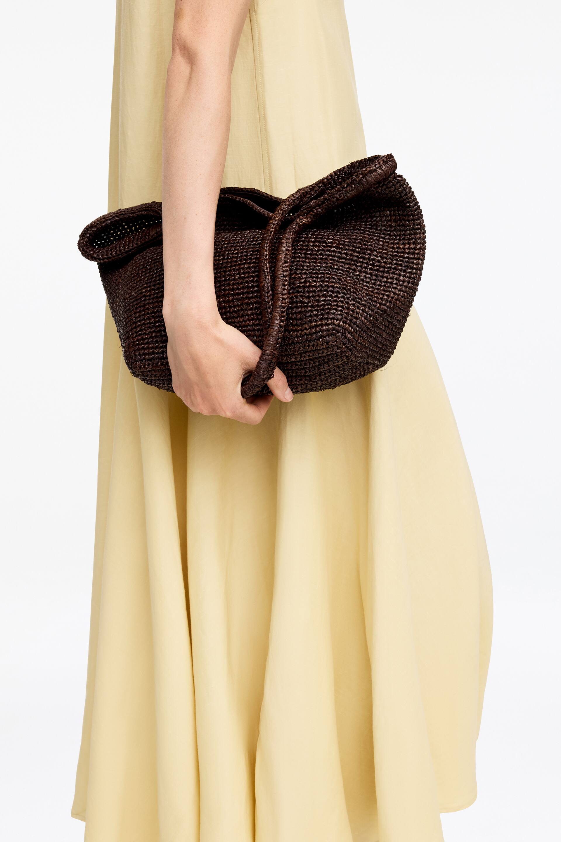 Größeres Bild anzeigen: Eine dunkelbraune geflochtene Tasche wird gehalten, die ein strukturiertes, kunstvolles Geflecht und eine geraffte obere Öffnung aufweist. Eine Hand greift den geflochtenen dunkelbraunen Kordelzuggriff.