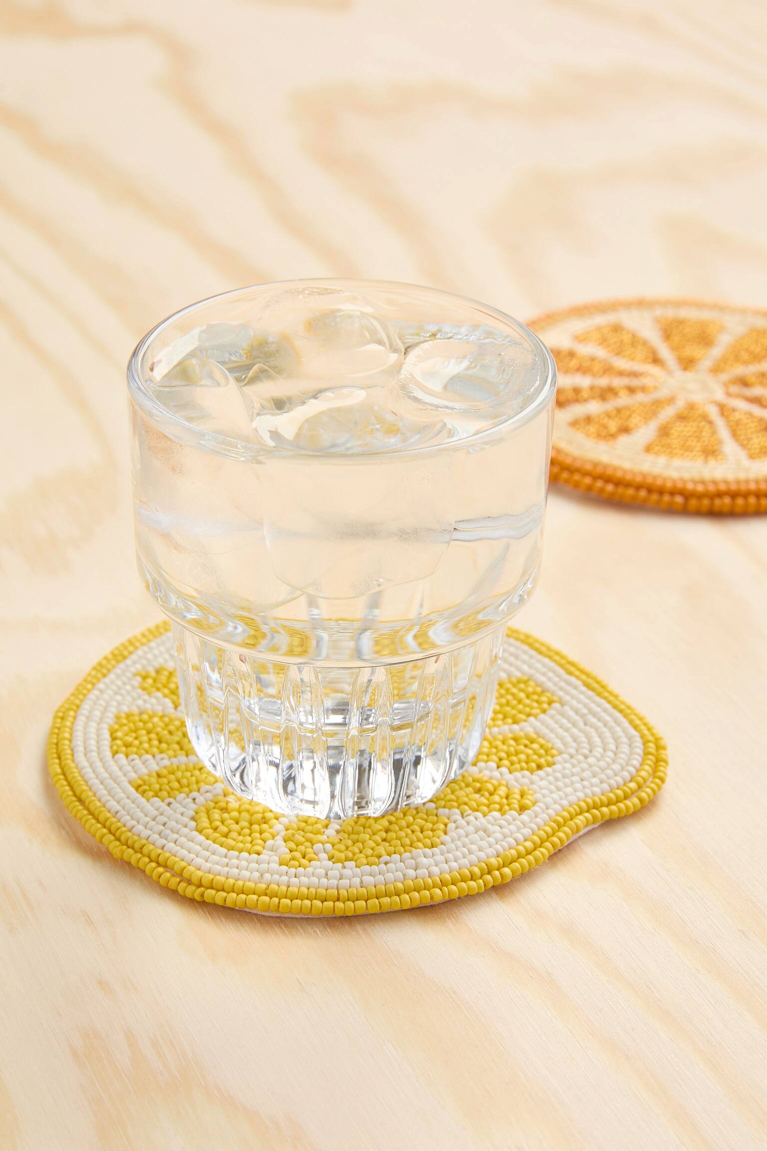 Un sottobicchiere rotondo, realizzato con perline di vetro gialle e bianche, presenta un motivo centrale a fette di limone. Ha un bordo leggermente irregolare.