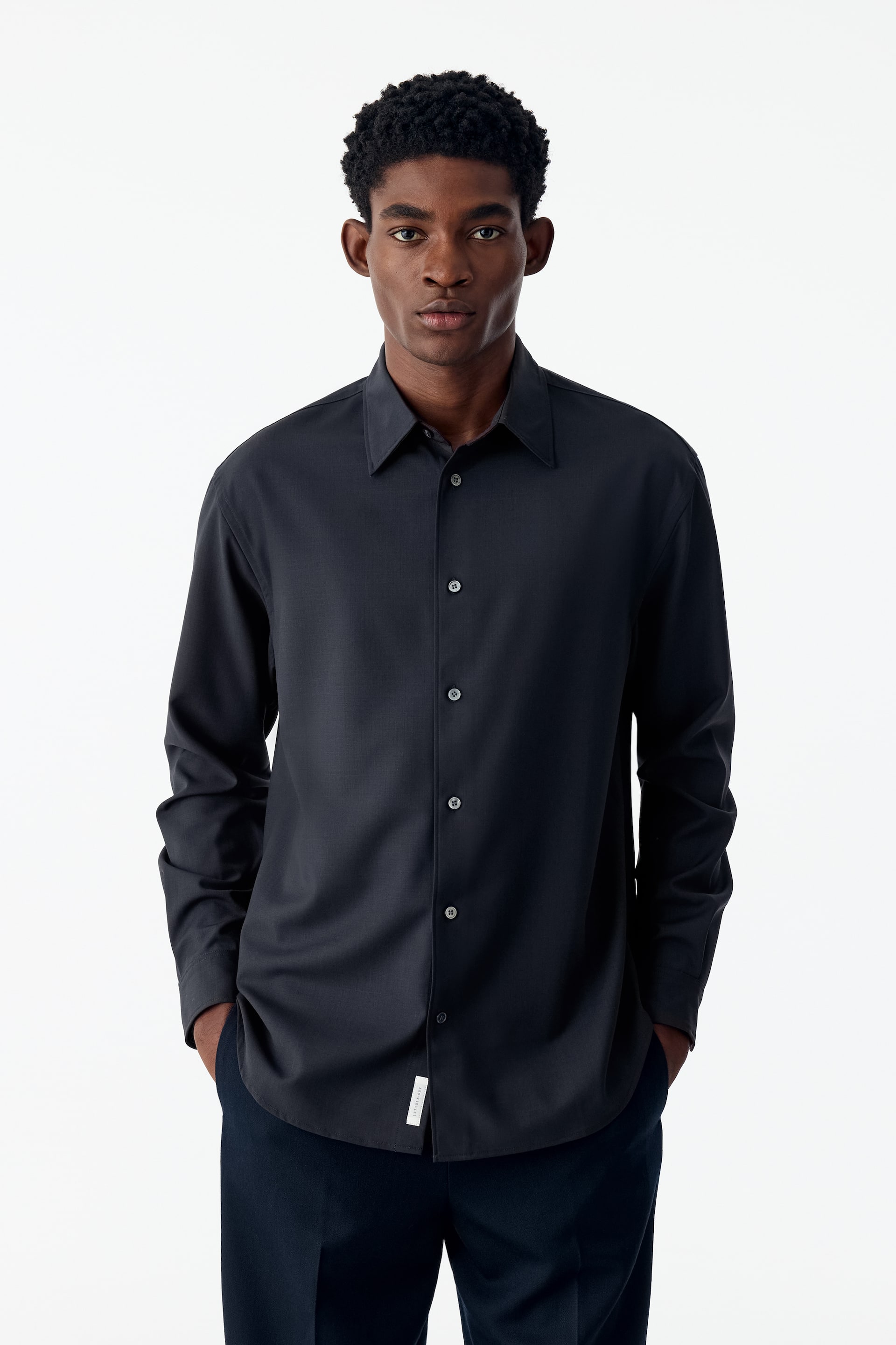 View larger image: The model is wearing a dark grey, long-sleeved button-up shirt with a relaxed fit, a point collar and a curved hem. The shirt is styled untucked over dark trousers, with the model’s hands resting in their pockets.