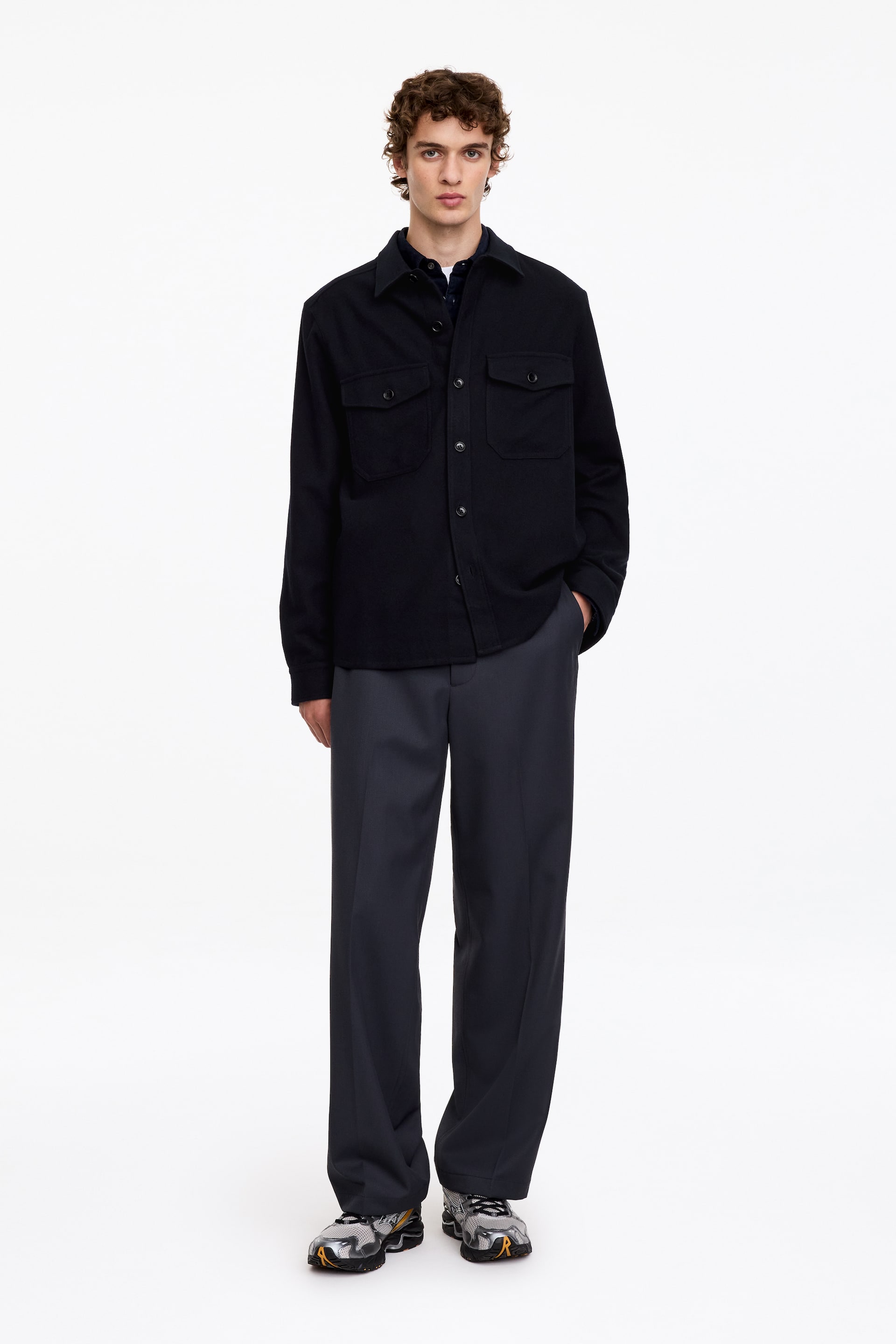View larger image: A black button-up overshirt featuring a pointed collar and two buttoned flap pockets on the chest is worn by the model. The overshirt is styled with dark grey wide-leg trousers and athletic trainers.