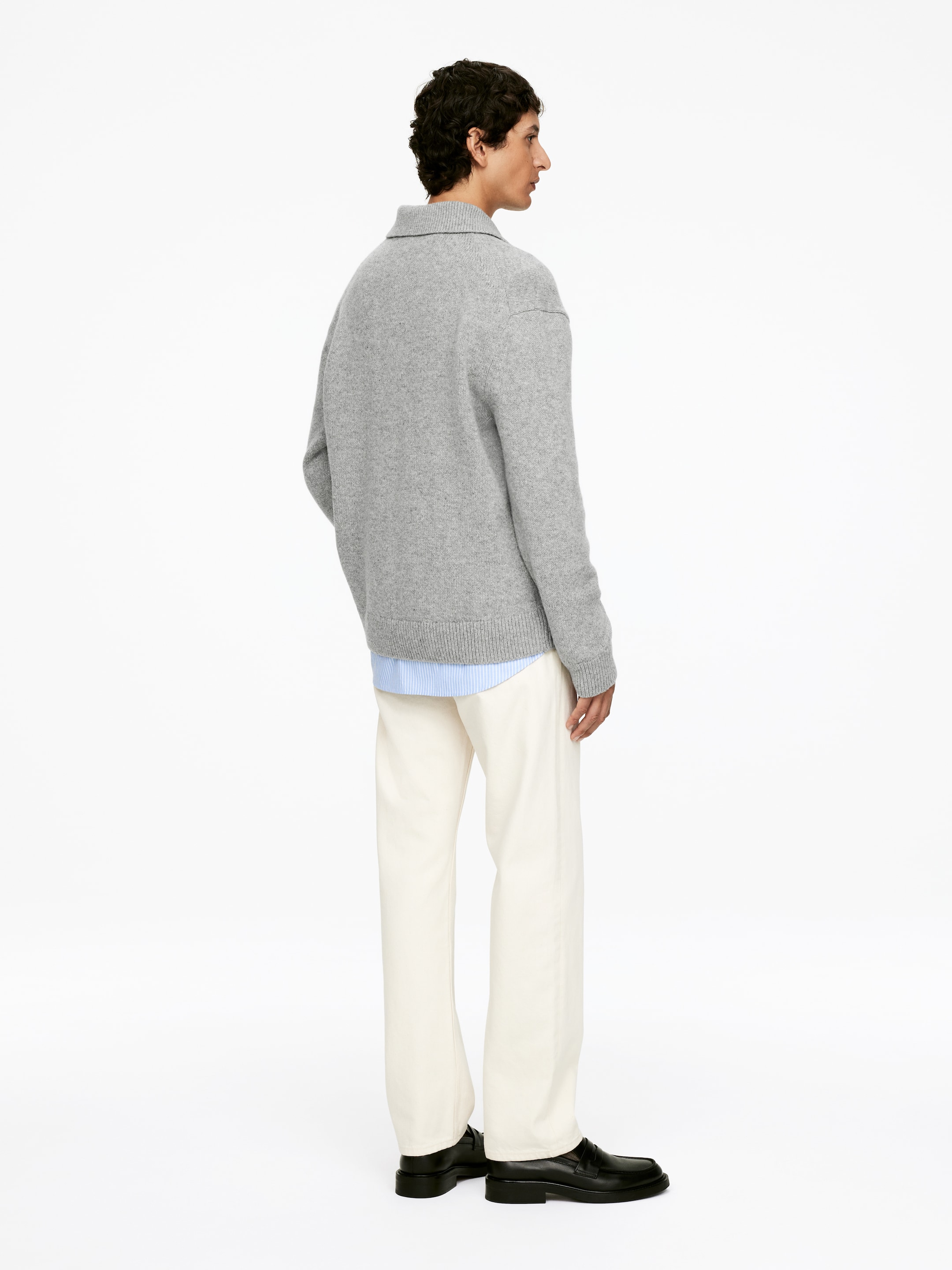 Größeres Bild anzeigen: Ein grauer Strickpullover wird von einer Person getragen, die nach hinten und leicht nach rechts schaut, und zeigt seine lockere Passform, den gerippten Saum und den weichen Kragen. Ein hellblau gestreiftes Hemd ist darunter geschichtet, sichtbar unter dem Saum des Pullovers, kombiniert mit cremefarbenen Hosen und schwarzen Loafers.