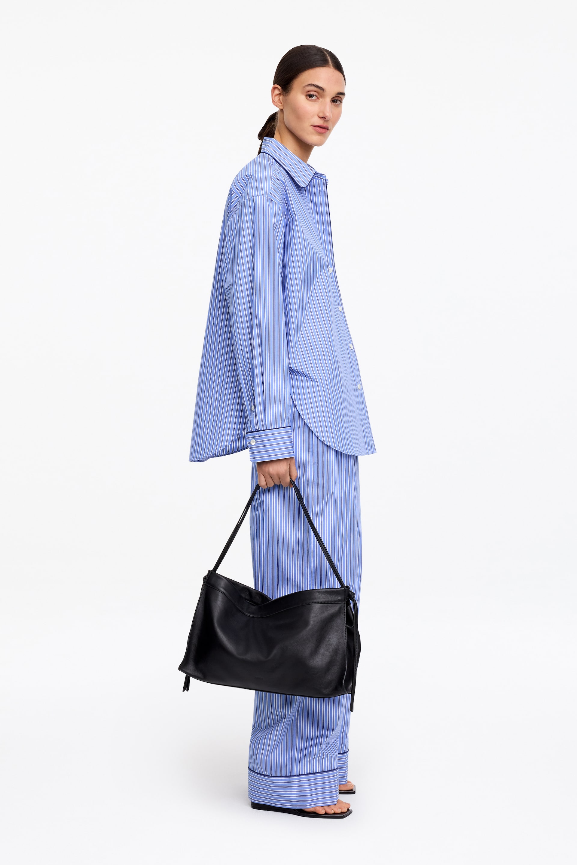 Ver imagen más grande: Un bolso de cuero negro holgado es sostenido por una mano, con una fina correa negra y detalles de cordón en sus lados. Muestra una forma rectangular suave con pliegues visibles, y su parte frontal está ligeramente orientada hacia la derecha.