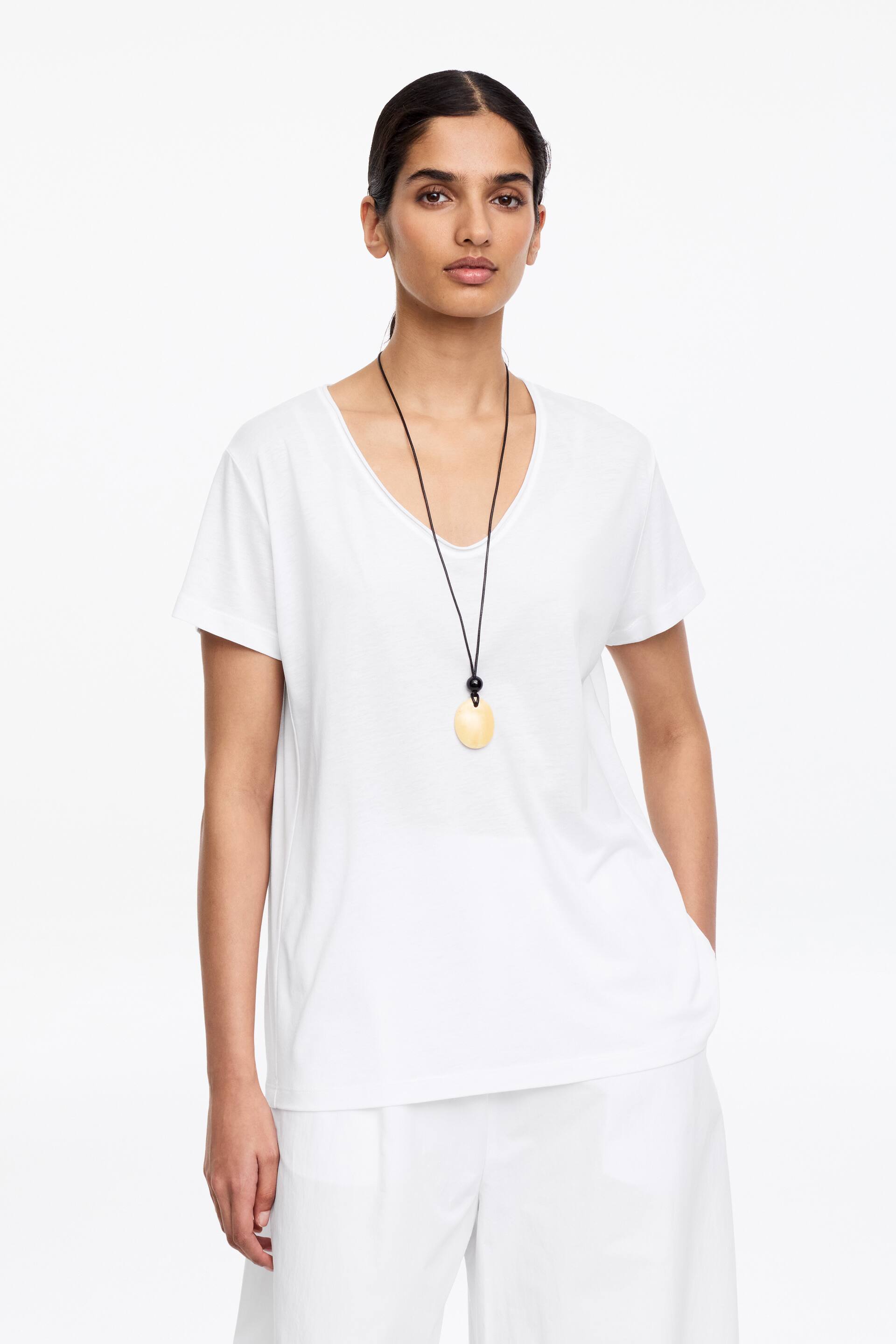 Se større billede: En hvid T-shirt med korte ærmer, en rund halsudskæring og rullekantdetaljer ved halsen og ærmerne bæres af modellen. T-shirten falder let og viser svagt buede sidesømme, der bevæger sig mod fronten, og er stylet med hvide shorts og en sort halskæde.