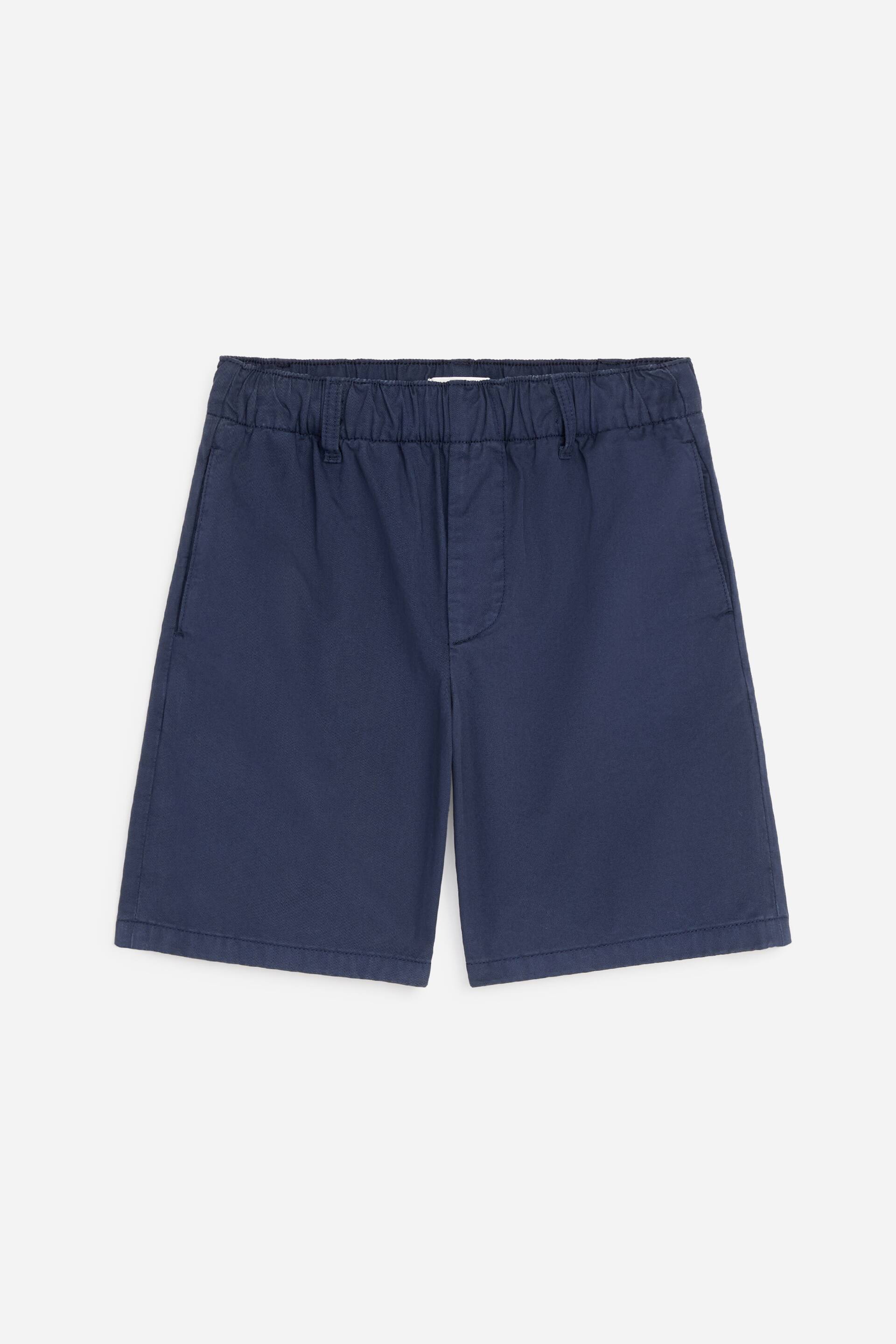 Visualizza immagine più grande: Shorts in twill di cotone blu scuro presentano il loro lato anteriore, caratterizzati da una vita elasticizzata con arricciature, tasche laterali a cucitura e un dettaglio di patta finta cucita.