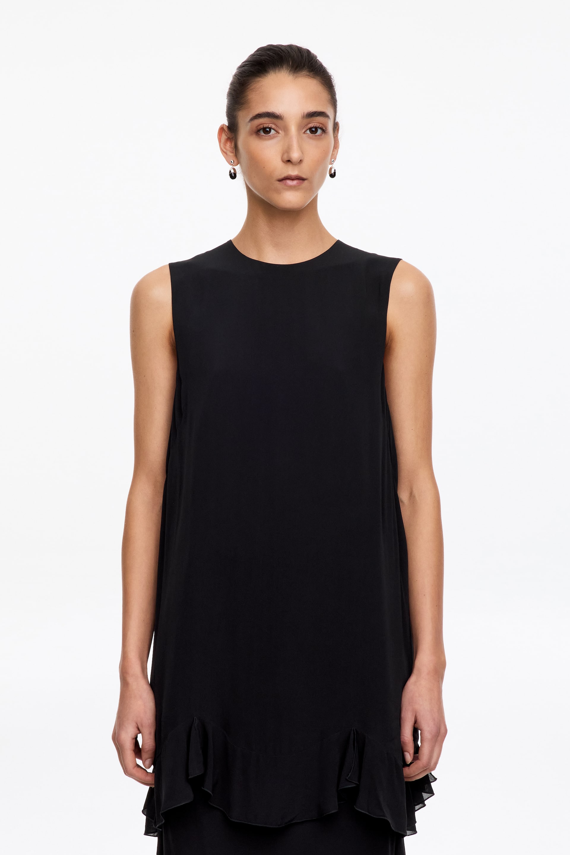 Grösseres Bild ansehen: Das Model trägt eine schwarze, ärmellose Tunika mit Rundhalsausschnitt und gewelltem, gerüschtem Saum. Das Regular-Fit-Kleidungsstück fällt locker und unterstreicht seine schlichte Silhouette sowie den weich fliessenden Saum.