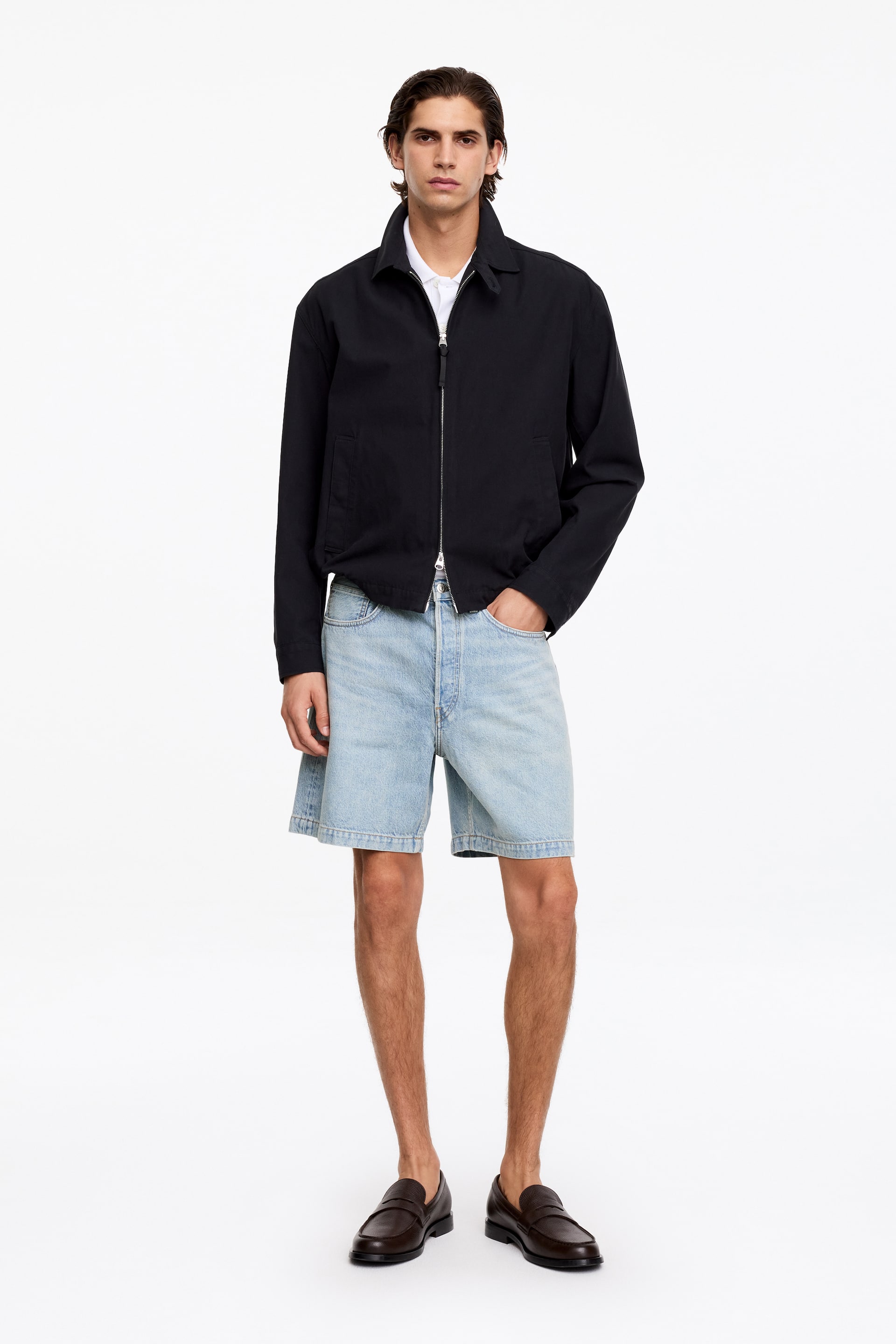 Grotere afbeelding bekijken: Een zwarte jas met een volledige ritssluiting aan de voorkant met een zilveren rits, een puntige kraag en twee zijzakken, gedragen over een witte poloshirt.