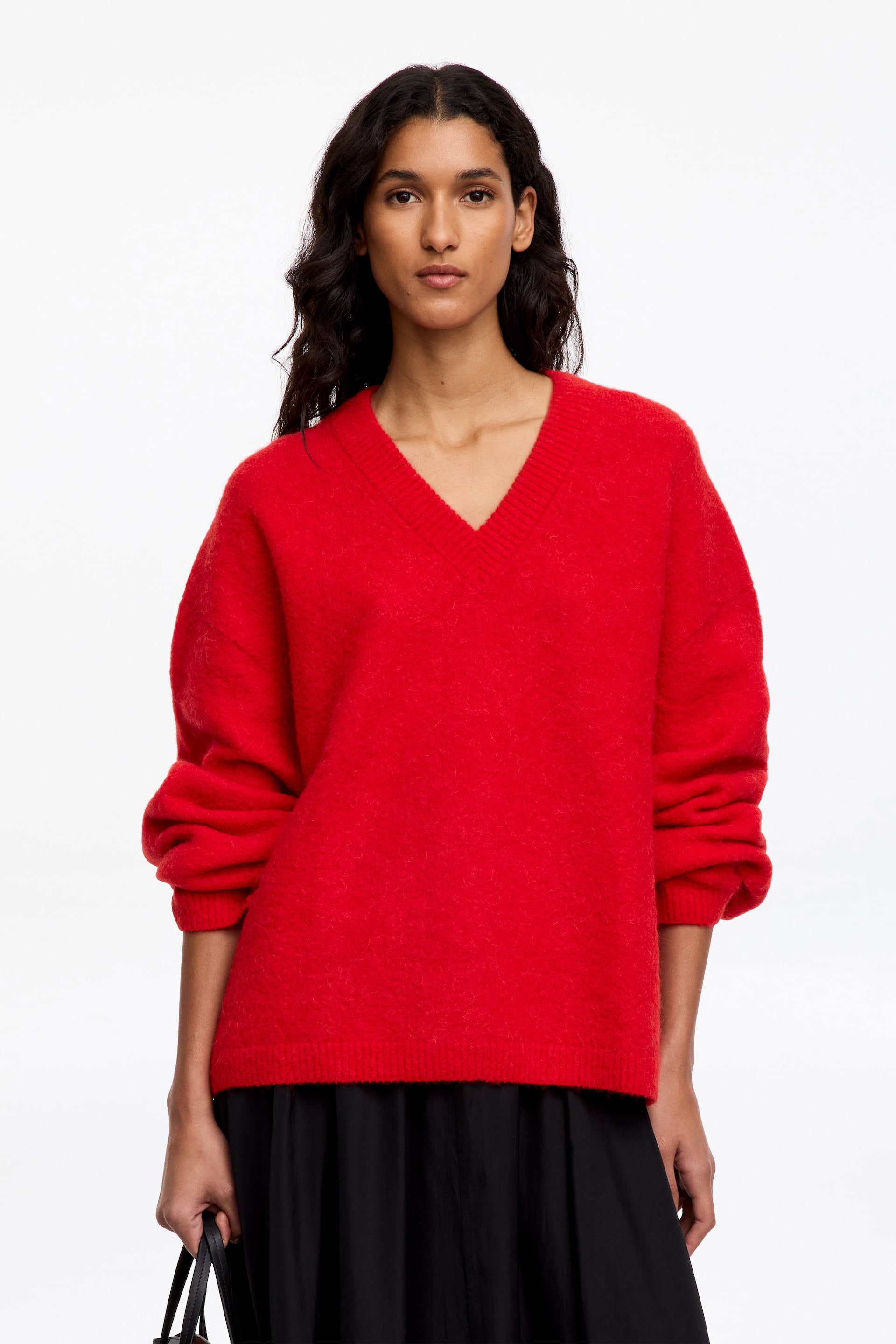 Ver imagem maior: A modelo está a usar uma camisola de malha vermelha com decote em V, corte descontraído e mangas compridas. A modelo também está a usar uma saia preta e a segurar uma mala preta.