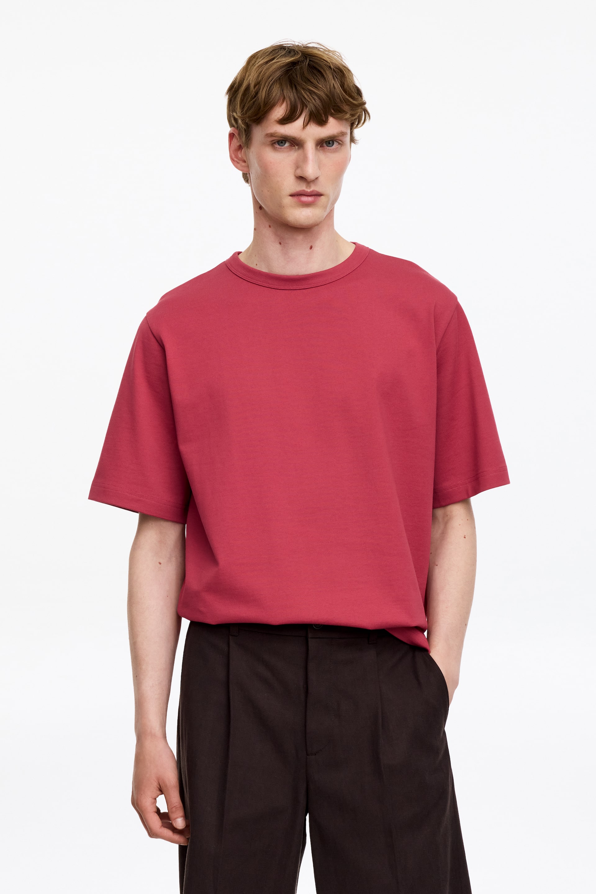 Grösseres Bild ansehen: Das rote T-Shirt verfügt über einen Rundhalsausschnitt und kurze Ärmel. Es sitzt locker und hängt ungesäumt über dunklen Hosen, wobei es aus einer frontalen Perspektive seinen entspannten Fall zeigt.