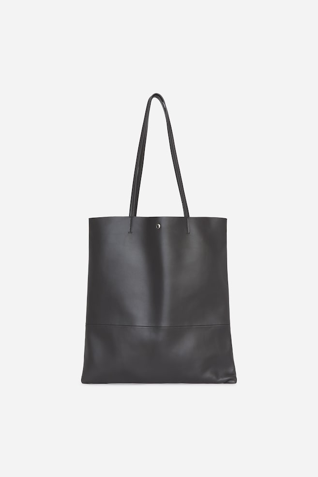 Un sac cabas en cuir gris foncé est présenté de face, mettant en valeur sa forme rectangulaire minimaliste et ses fines anses bandoulière. Une petite fermeture à bouton-pression argenté est visible au centre supérieur, et une couture horizontale nette traverse la partie inférieure de la surface lisse du sac. - 1