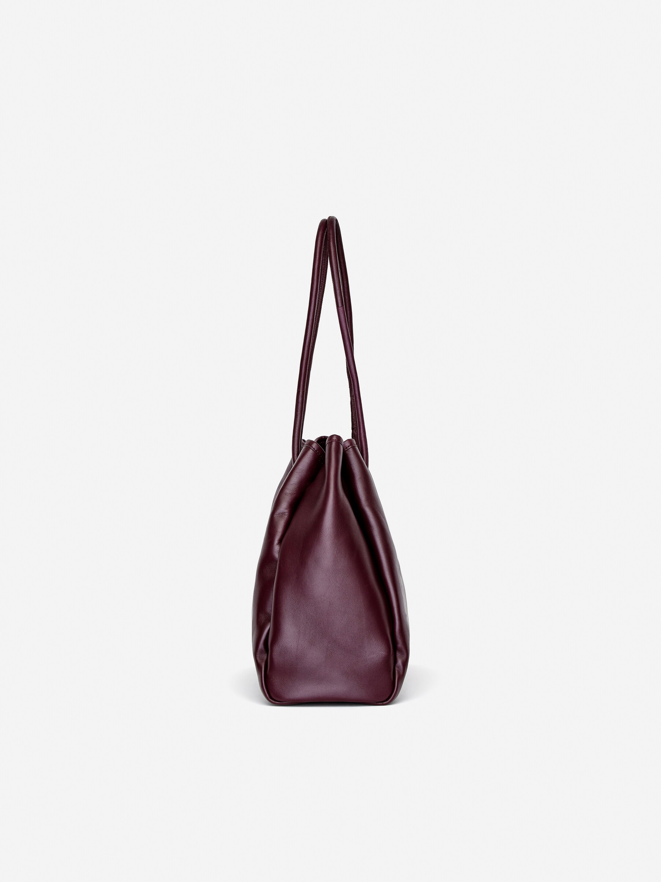 Agrandir l'image: Un sac fourre-tout en cuir est debout, légèrement incliné vers la gauche. Il est fabriqué en cuir bordeaux foncé et doux, avec une ouverture supérieure froncée et deux poignées supérieures fines formant une forme de V.