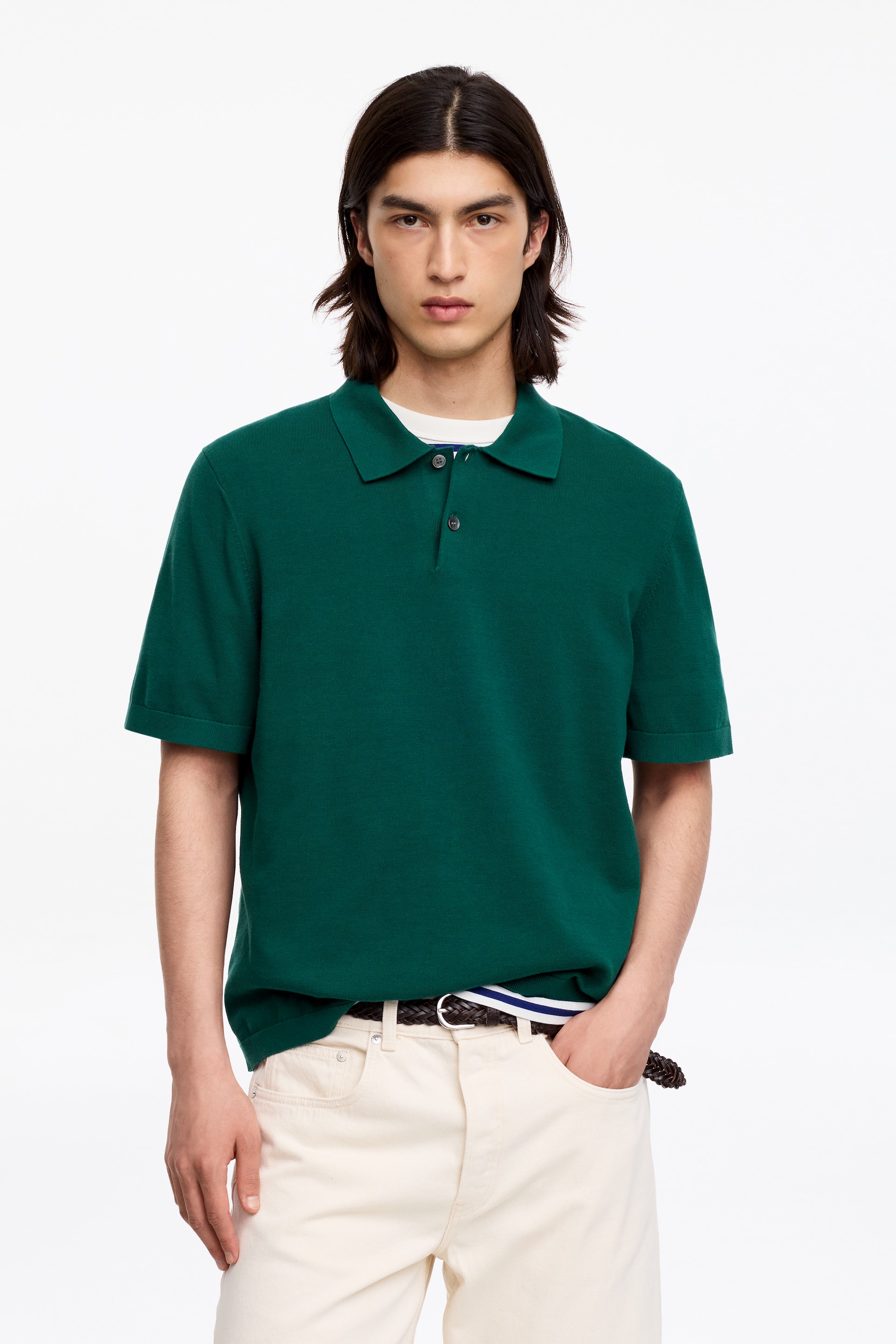 Grotere afbeelding bekijken: Een donkergroen gebreid poloshirt, met korte mouwen en een tweeknoopssluiting met donkere knopen, wordt gedragen door het model. Een wit T-shirt is eronder gelaagd, zichtbaar bij de taille, en crèmekleurige broek wordt gedragen met een gevlochten bruine riem.