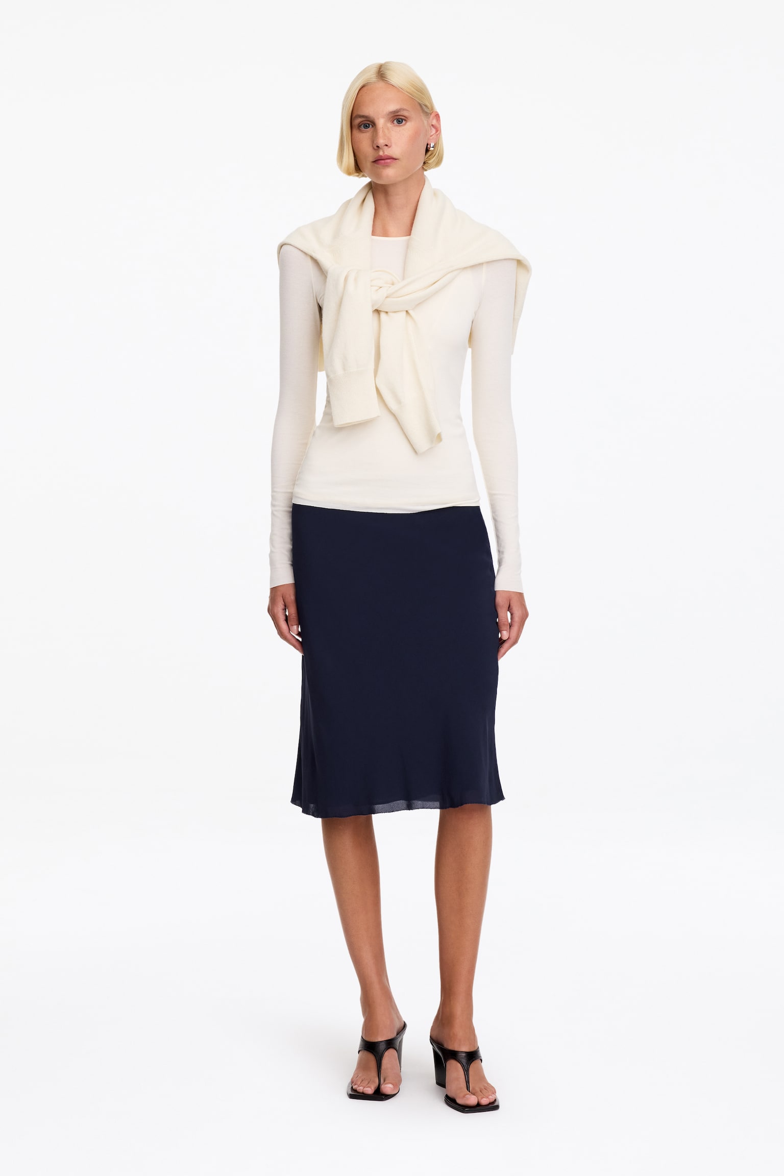Une jupe midi en mousseline bleu marine est portée par le mannequin, mettant en valeur une silhouette droite et un ourlet double couche subtilement transparent. La jupe est associée à un haut crème à manches longues, à un pull crème assorti posé sur les épaules, ainsi quâà des sandales noires à talons. - 1