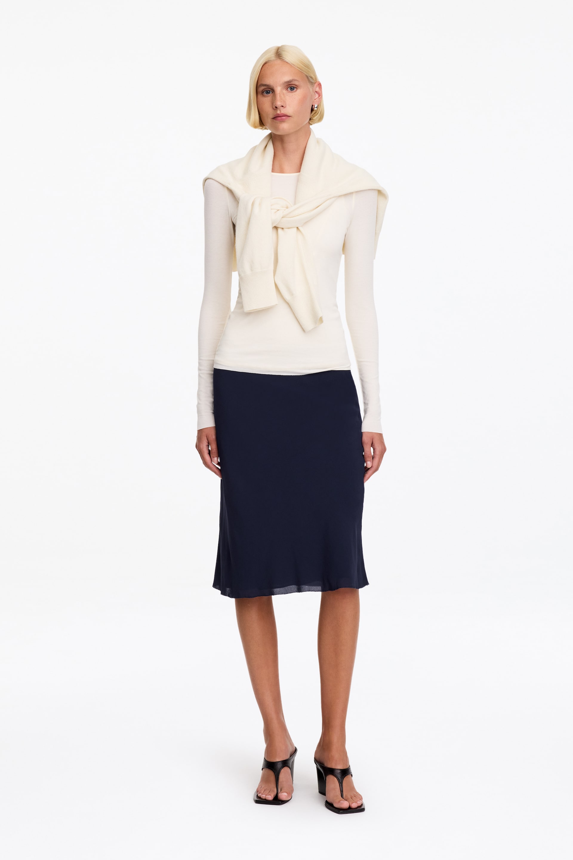 View larger image: The model is wearing a navy blue chiffon midi skirt, showcasing a straight silhouette and a subtly sheer double-layer hem. The skirt is styled with a long-sleeved cream top, a matching cream sweater draped over the shoulders, and black heeled sandals.