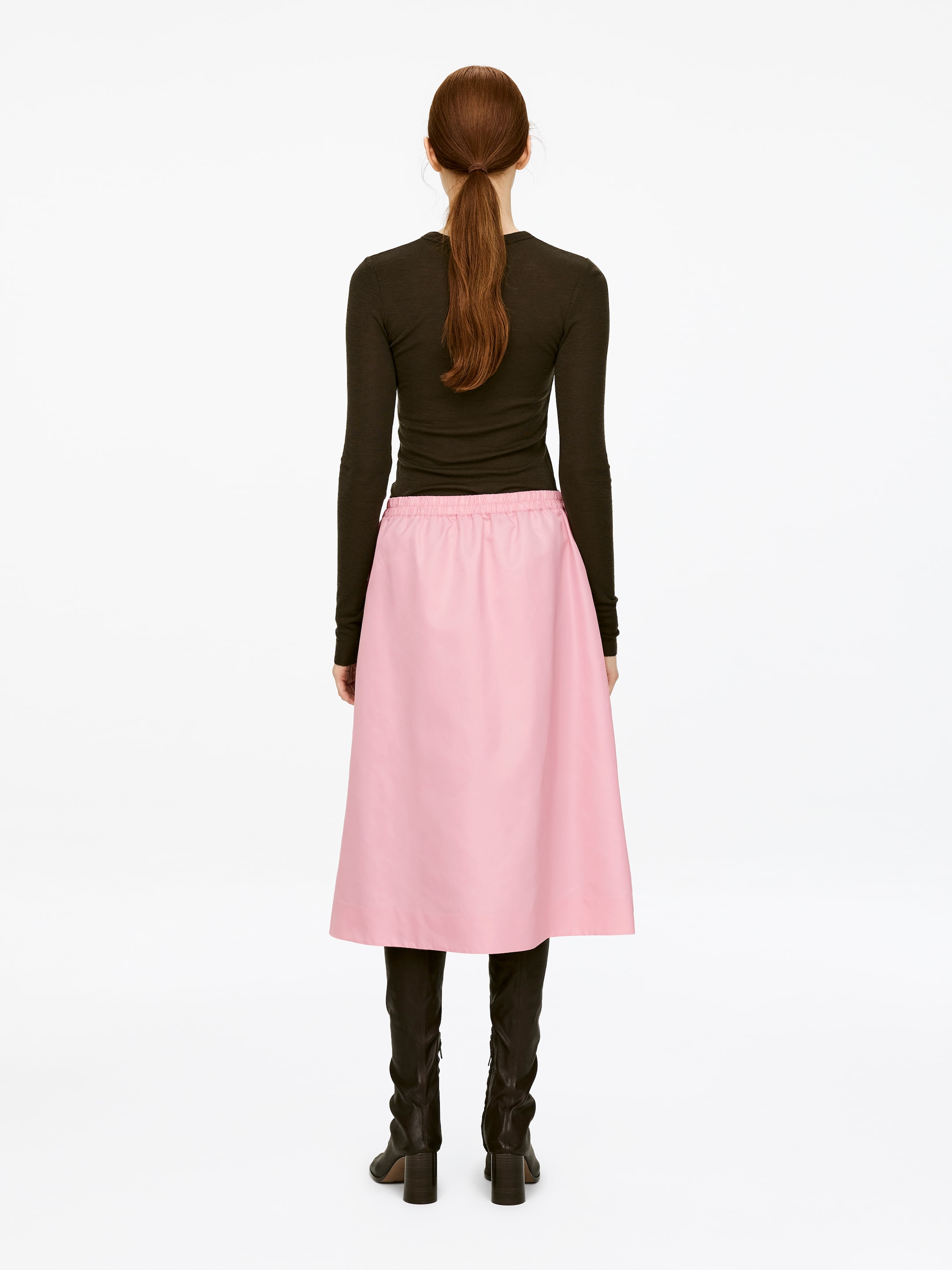 View larger image: A pink midi skirt featuring an elasticated waist is being worn by the model, who is also wearing a dark long-sleeved top and black boots.