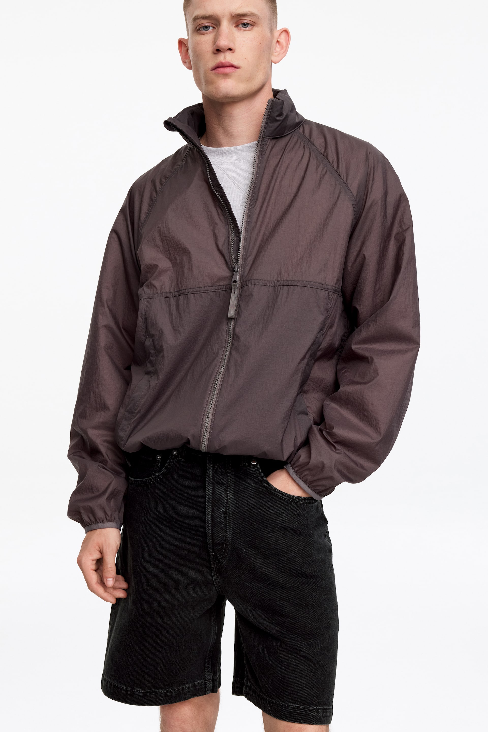Agrandir l'image: Une veste semi-transparente d'un brun foncé atténué est présentée, avec un col montant et une fermeture éclair frontale sur toute la longueur. Elle est conçue avec des manches raglan et des poignets et un ourlet élastiqués, créant une coupe décontractée sur un t-shirt gris clair visible.