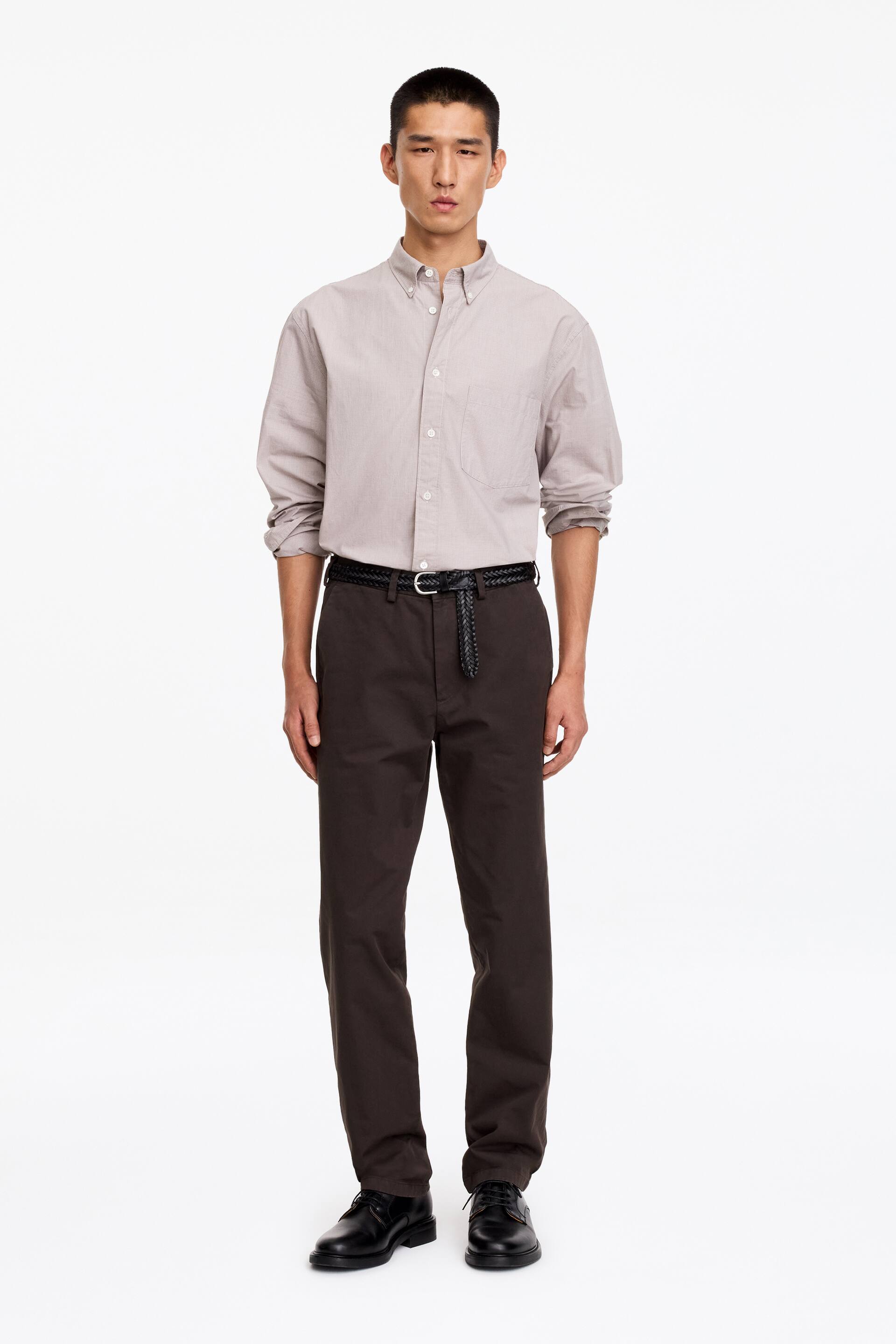 Agrandir l'image: Un chino en coton marron foncé, coupe régulière et droite, est porté par le mannequin avec une ceinture tressée noire passée dans les passants. Le chino est associé à une chemise boutonnée gris clair rentrée dans le pantalon et à des chaussures en cuir noires.