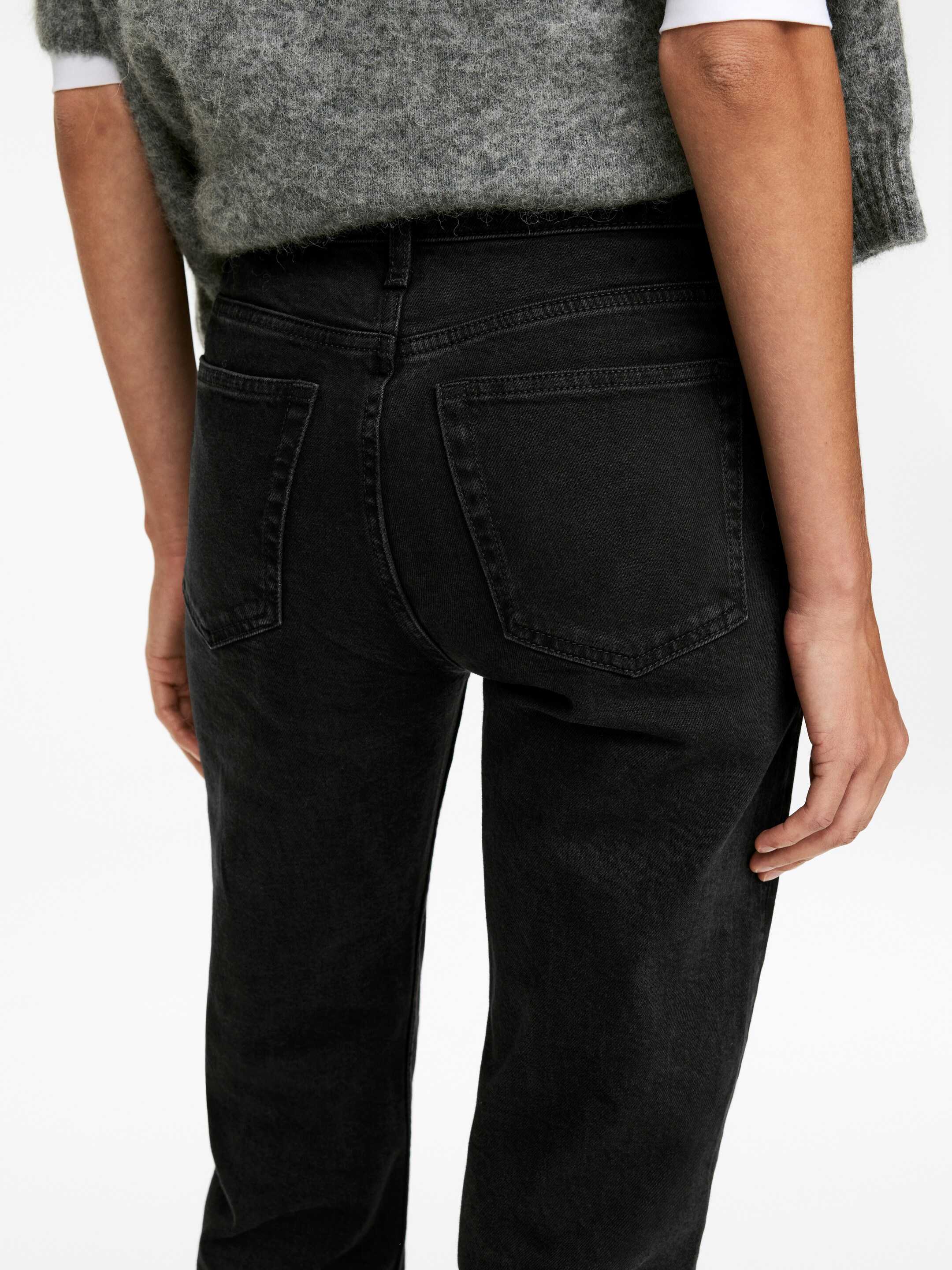 Visualizza immagine più grande: Un paio di jeans in denim nero viene mostrato da dietro, leggermente inclinato a sinistra, mettendo in evidenza la sua vestibilità a vita alta. I jeans presentano un lavaggio nero sbiadito, un carré posteriore a forma di V e due tasche posteriori quadrate con cuciture visibili.