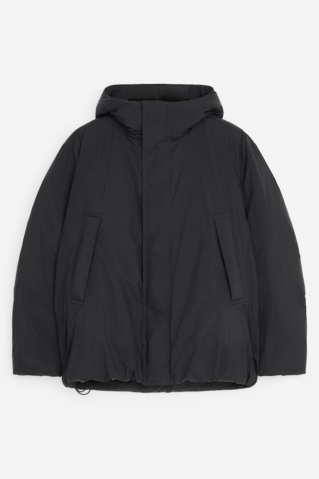 Una chaqueta acolchada negra con capucha está de frente, mostrando un tejido de textura mate, una capucha acolchada y un cierre frontal oculto. La chaqueta presenta dos grandes bolsillos de vivo inclinados y un bajo ajustable con cordón. - 1
