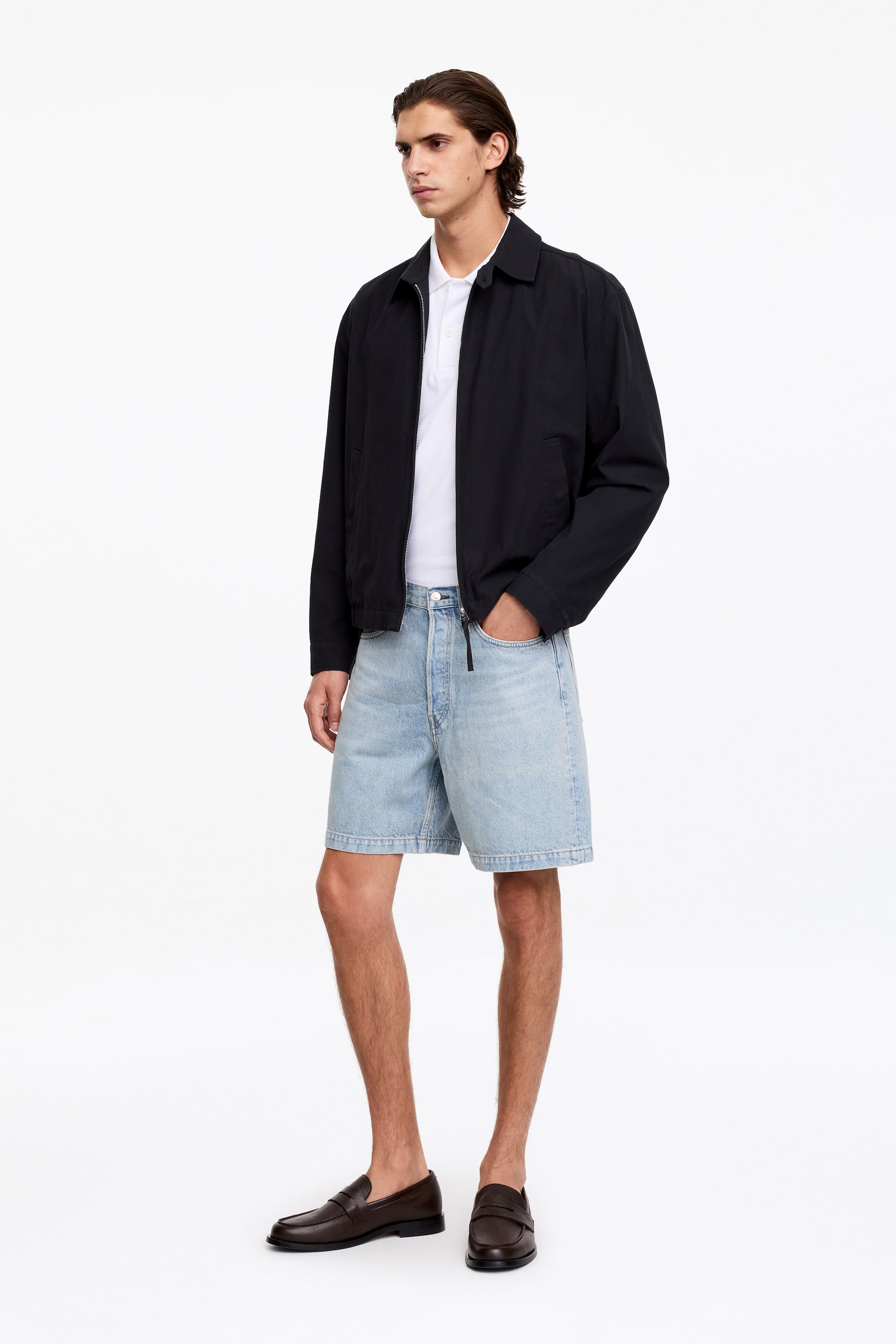 Agrandir l'image: Short en denim à délavage clair avec 5 poches, coupe décontractée et braguette à boutons apparente, portés par le mannequin. Le mannequin glisse une main dans une poche avant du short.