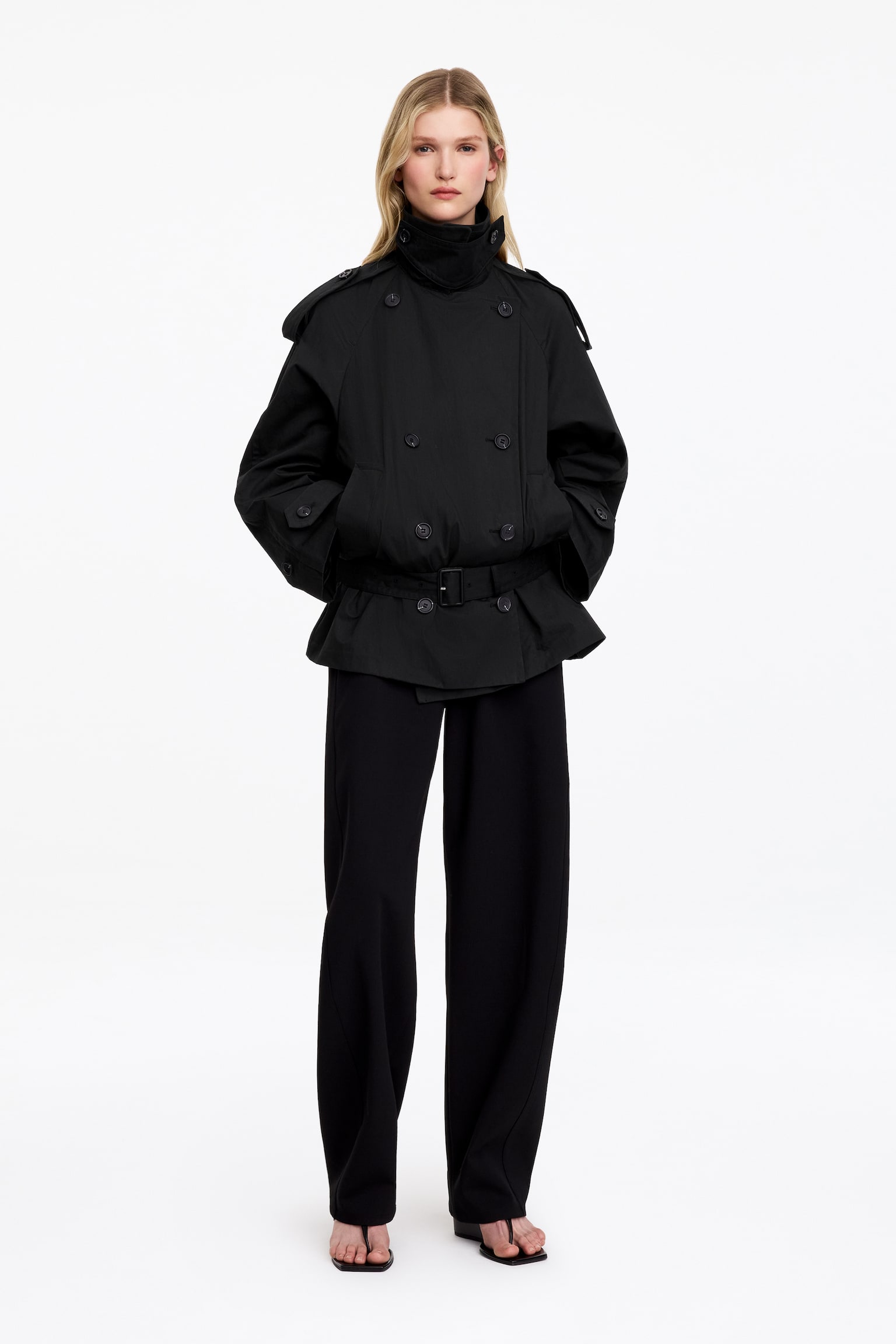Een zwarte trenchcoat wordt gepresenteerd VOOR, met een dubbele rij knopen, een puntige kraag met stormflap en schouderepauletten. Een stoffen riem met een rechthoekige gesp accentueert de taille, en de lange mouwen eindigen met manchetbandjes met knopen. - 1