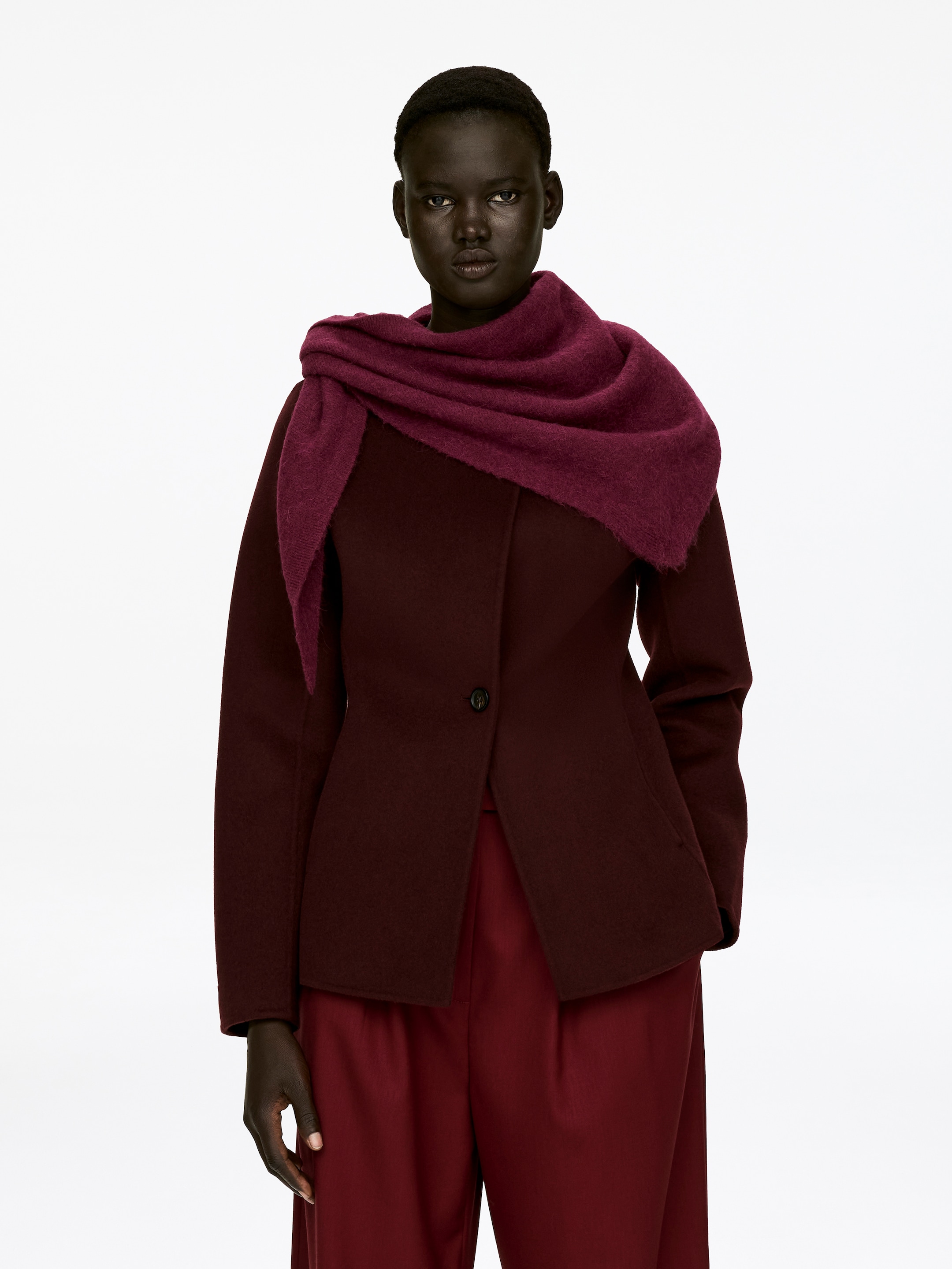 Visualizza immagine più grande: Una sciarpa in misto lana d'alpaca di un intenso color magenta è drappeggiata intorno al collo e alle spalle della modella, esibendo una texture morbida e pelosa. È stratificata sopra una giacca di un borgogna scuro.