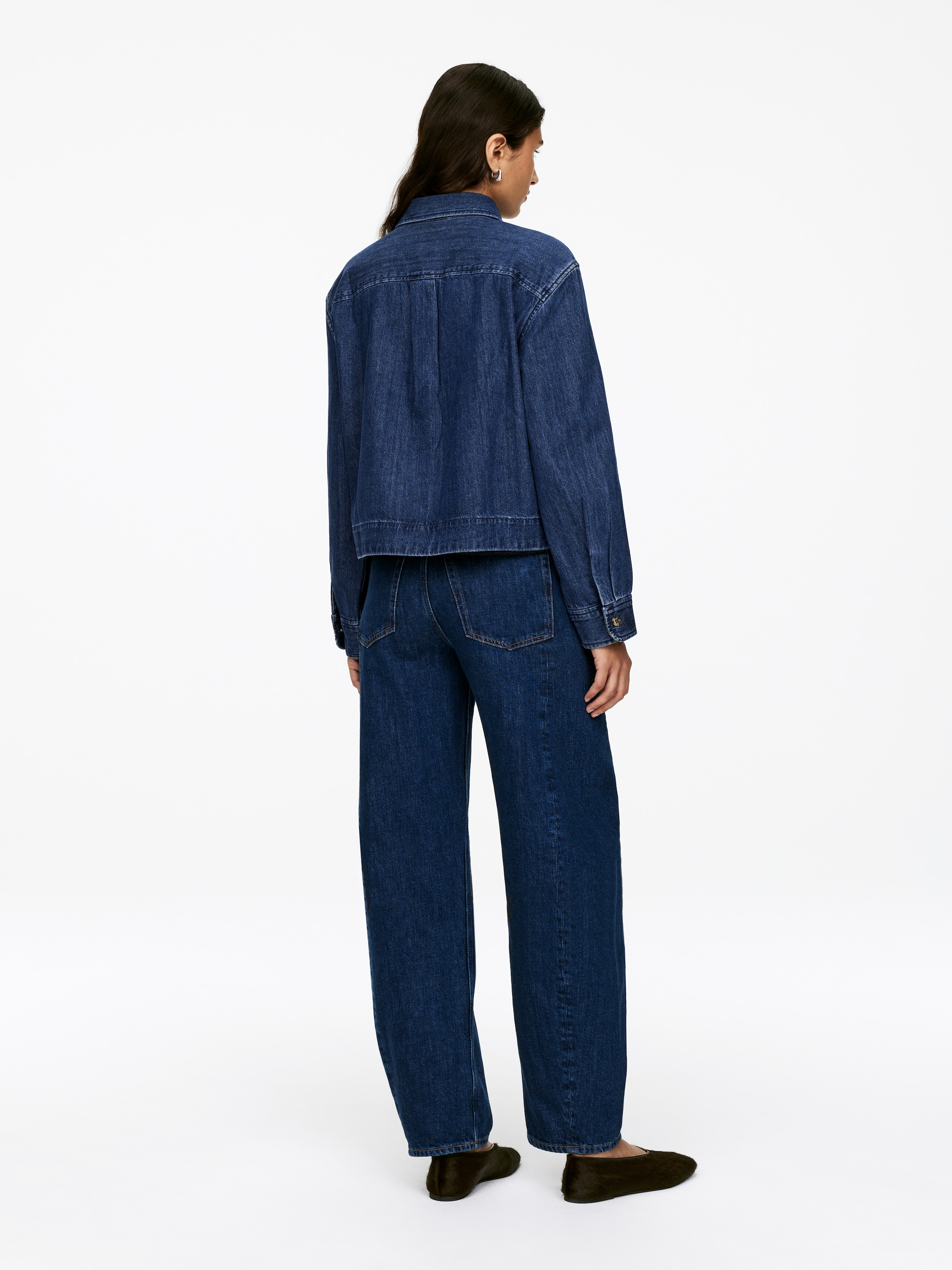 Visualizza immagine più grande: Una camicia di jeans blu scuro, caratterizzata da una silhouette corta e una vestibilità regolare, è indossata dalla modella. La modella indossa anche pantaloni di jeans blu scuro a gamba larga abbinati.