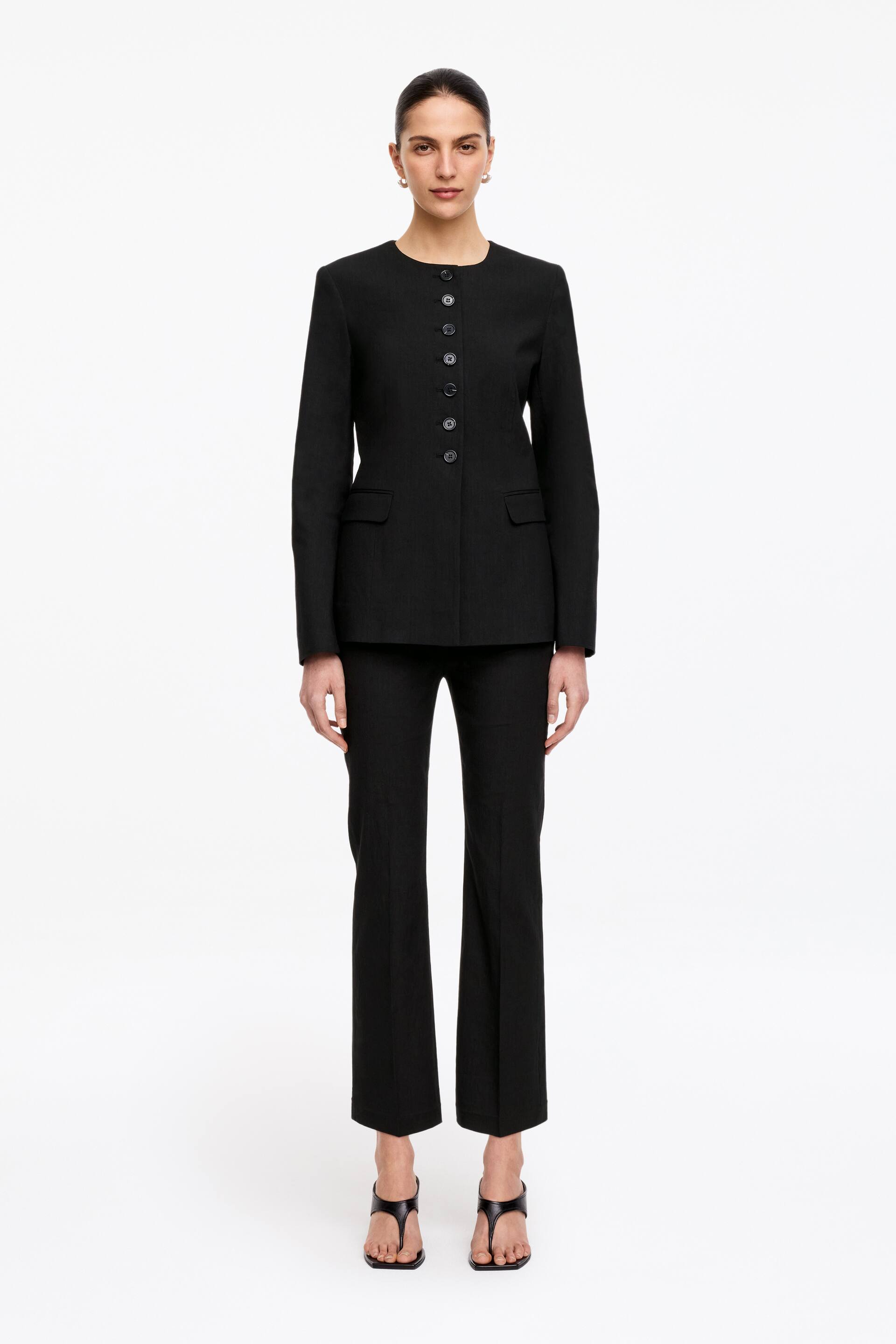 Ingrandisci l'immagine: Un tailleur pantalone nero viene presentato frontalmente, caratterizzato da una giacca senza collo con scollo rotondo, maniche lunghe, chiusura frontale con sei bottoni e due tasche con patta. I pantaloni abbinati sono a gamba dritta e corti alla caviglia.