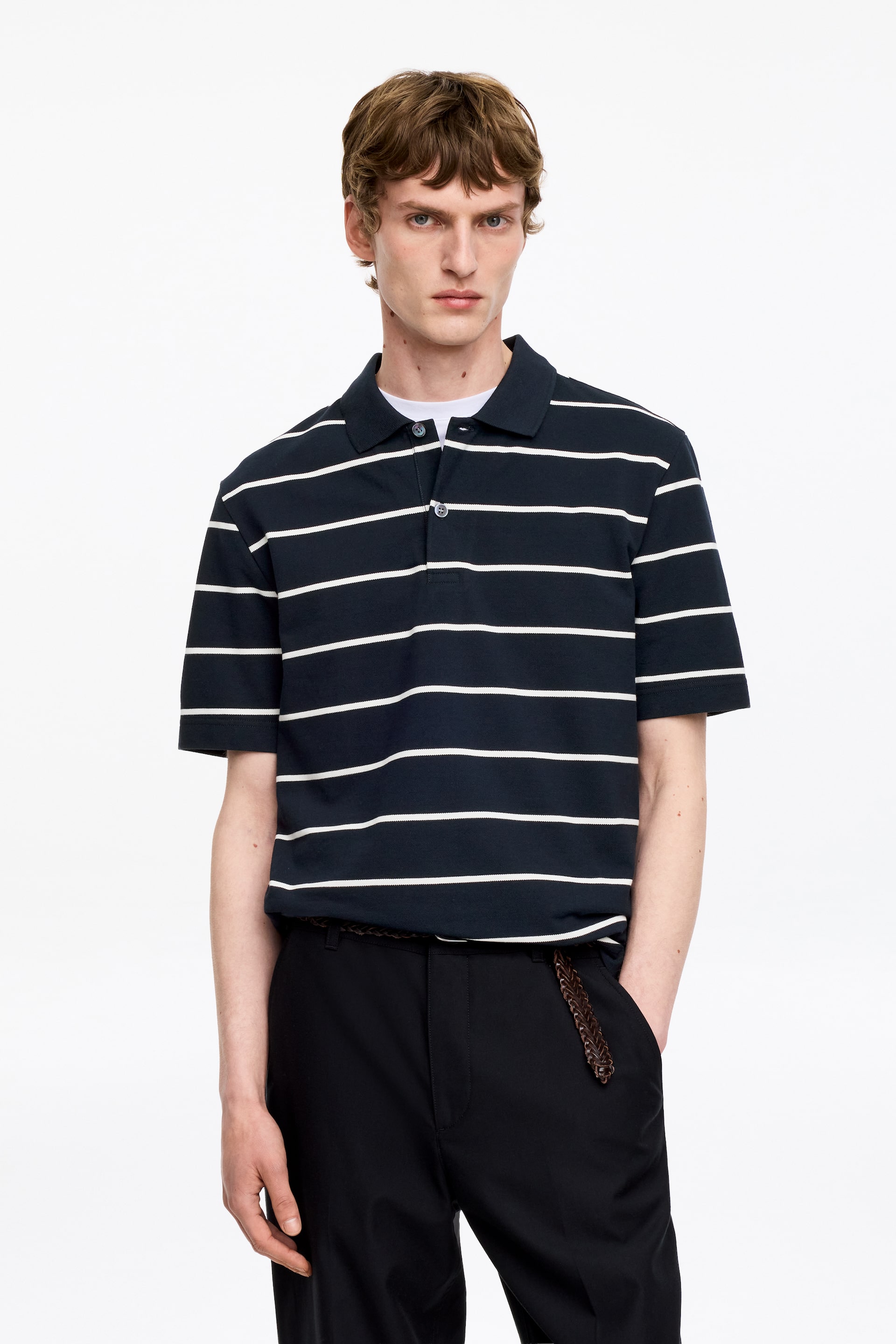 Größeres Bild ansehen: Ein marineblaues Poloshirt zeigt unterschiedliche horizontale weiße Streifen. Es verfügt über einen klassischen Kragen und eine Zwei-Knopf-Leiste, wobei ein weißes T-Shirt sichtbar unter dem Ausschnitt liegt.
