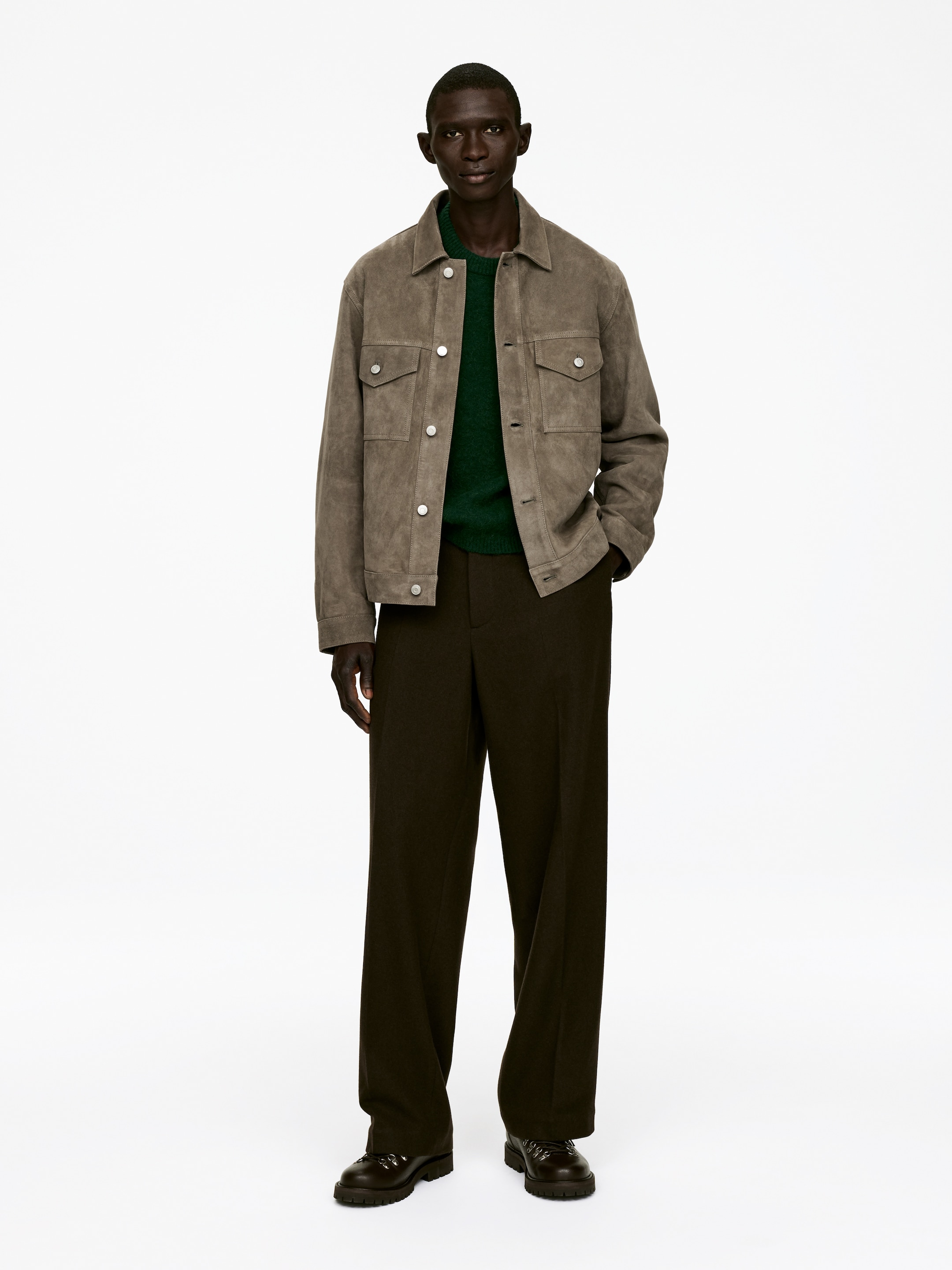 Agrandir l'image: Une veste en daim de couleur marron clair, avec une texture suédée, dotée d'un col classique, de deux poches poitrine avec rabats boutonnés et de deux poches passepoilées inférieures, est superposée sur un pull à col rond vert foncé. Elle est associée à un pantalon large de couleur marron foncé et des bottes à lacets sombres avec une semelle épaisse et crantée, présentée en vue frontale de plein corps.