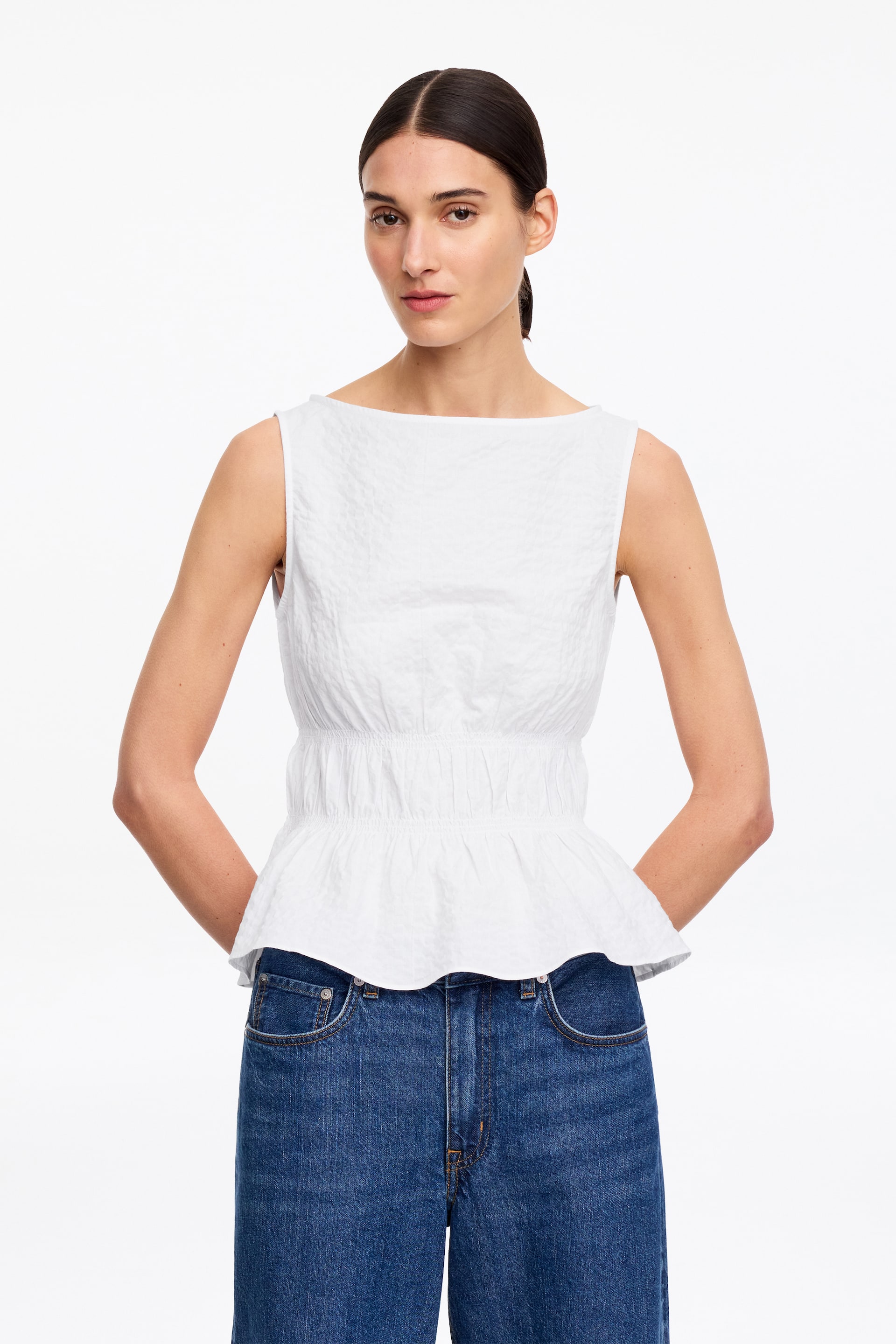 View larger image: The model is wearing a white seersucker top with a wide neckline and a smocked waist. The top is paired with blue denim jeans.