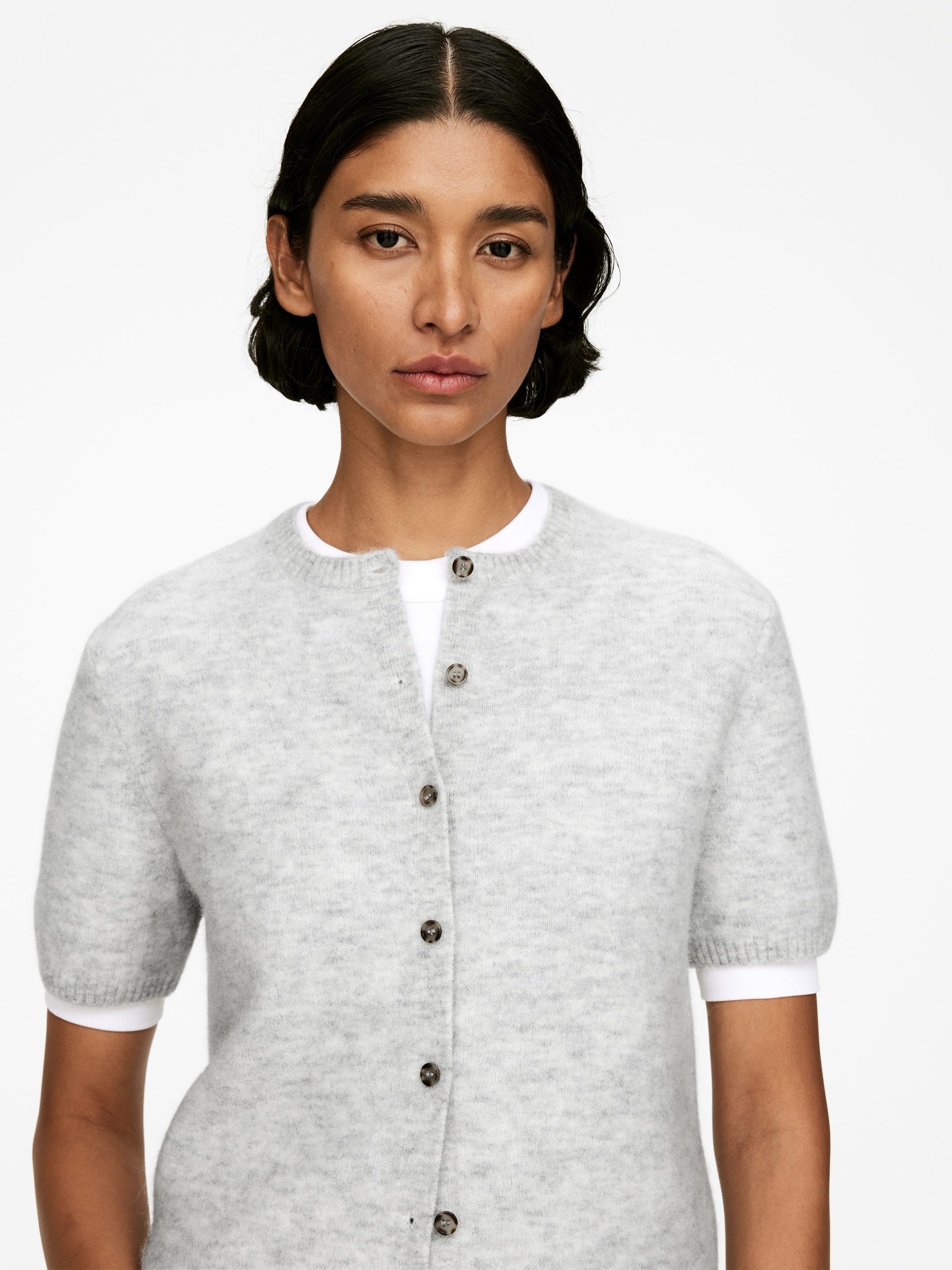 Grotere afbeelding bekijken: De model draagt een lichtgrijs gebreid vest met korte mouwen en een ronde hals, met donkere knopen langs de voorste knopenlijst, over een wit t-shirt.