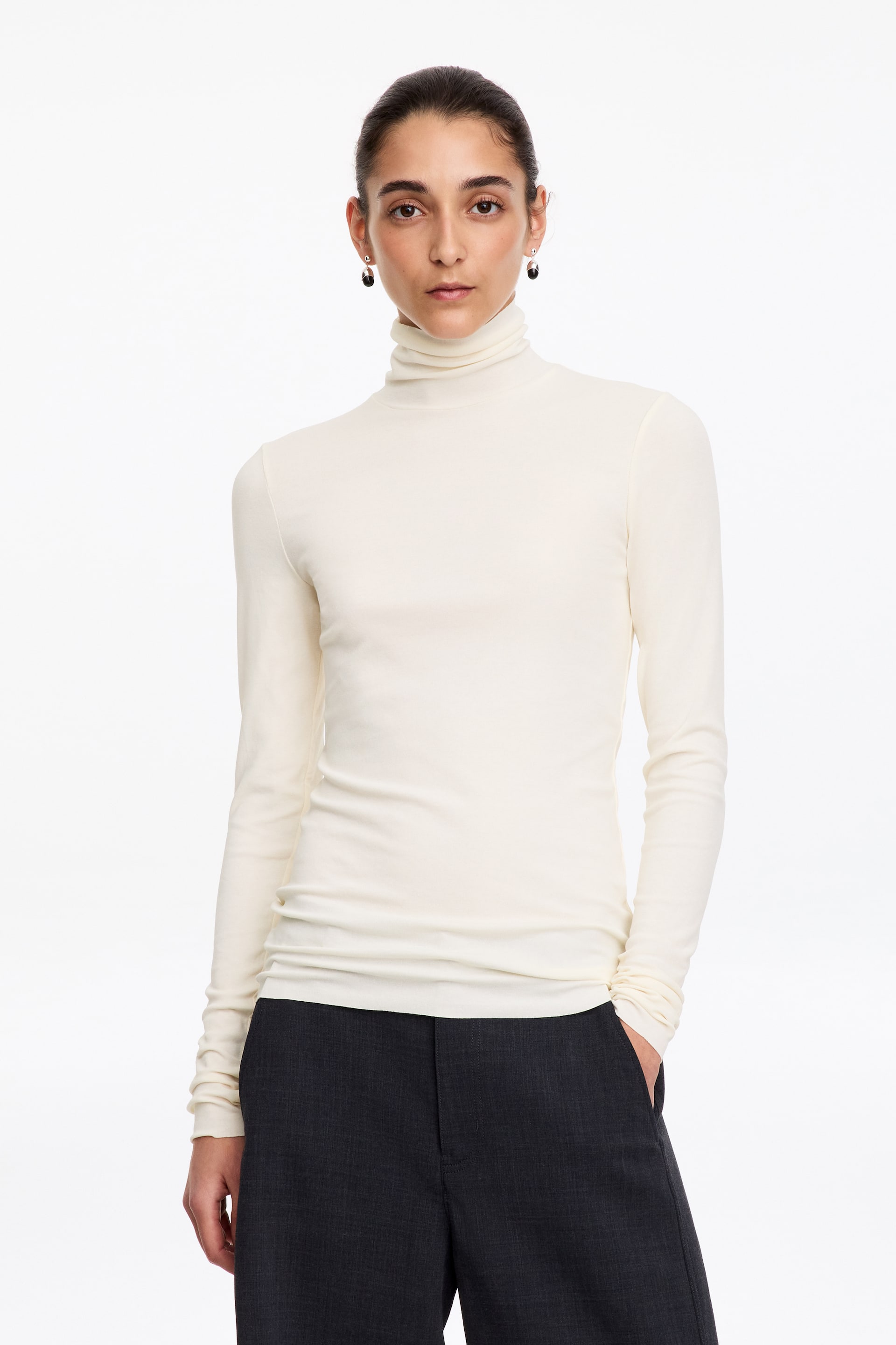 Größeres Bild anzeigen: Das Model trägt ein cremefarbenes Jersey-Top mit langen Ärmeln und Rollkragen in semitransparenter Optik. Das taillierte Oberteil ist mit dunkelgrauer Hose kombiniert.